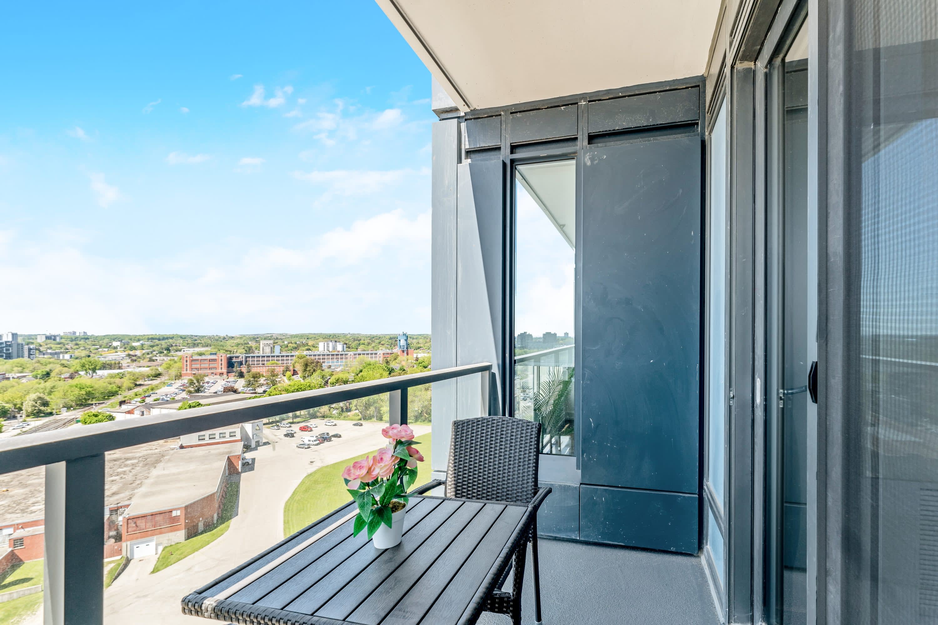 Balcony with a black wicker table and chair, featuring a pink floral centerpiece, overlooking a city landscape with buildings and a parking lot under a clear blue sky.