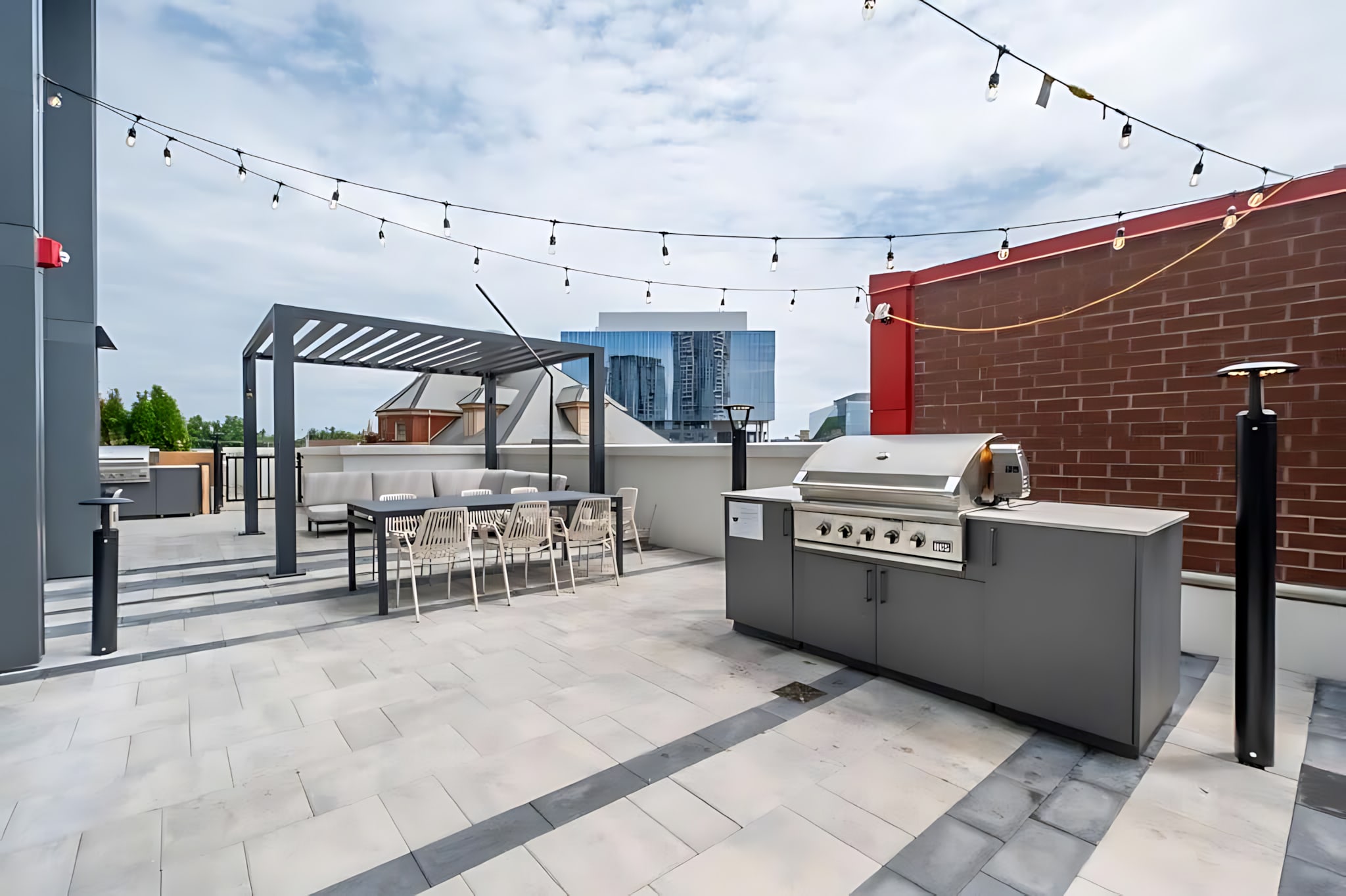 A modern rooftop patio features a grey pergola over a dining table and lounge seating, alongside a built-in BBQ station, all under string lights with city buildings in the background.