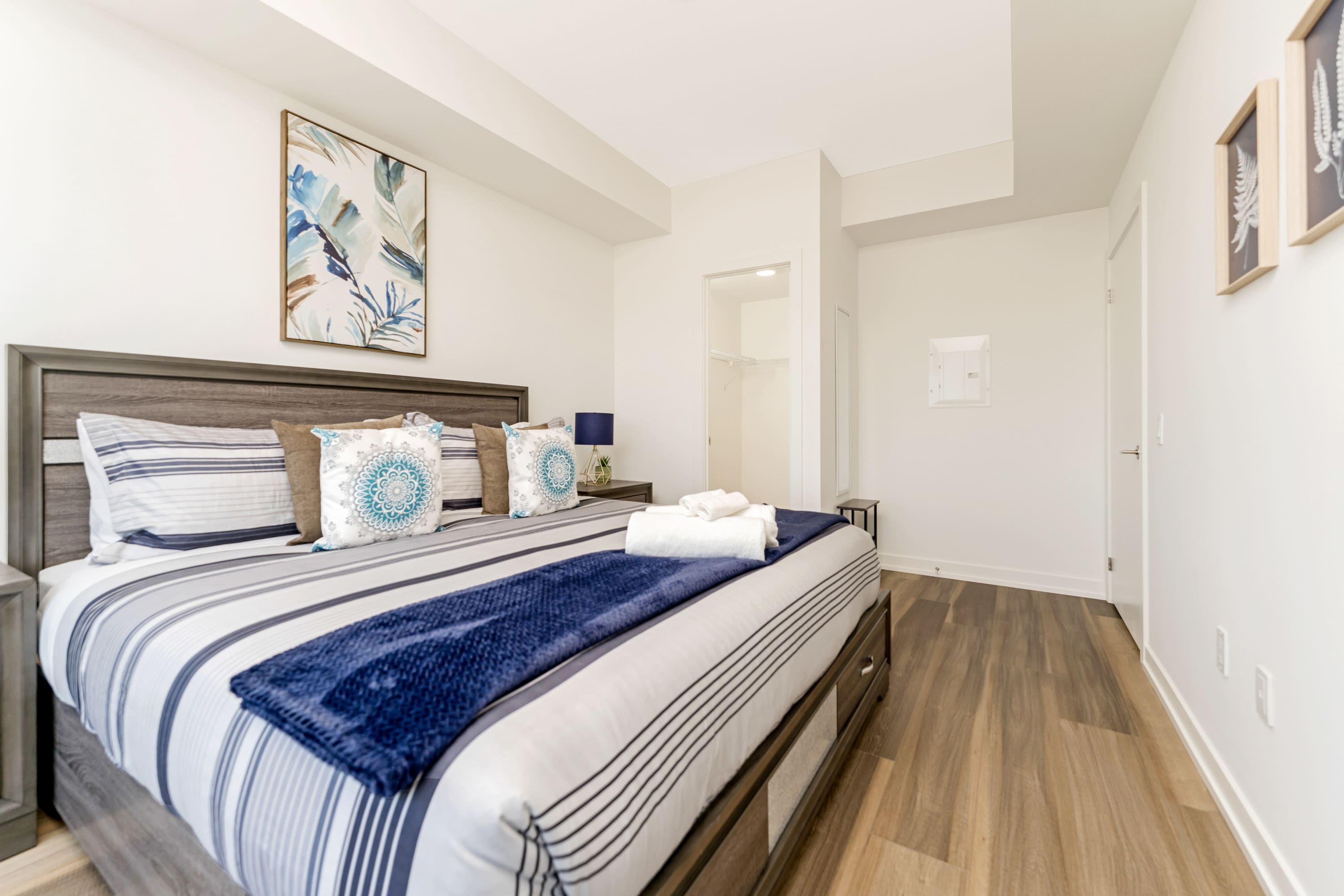 A spacious bedroom with a dark wood bed frame and headboard, featuring a striped comforter with blue and white patterns, accented by decorative pillows and a dark blue throw blanket. Light wood-look flooring and white walls complete the modern look.
