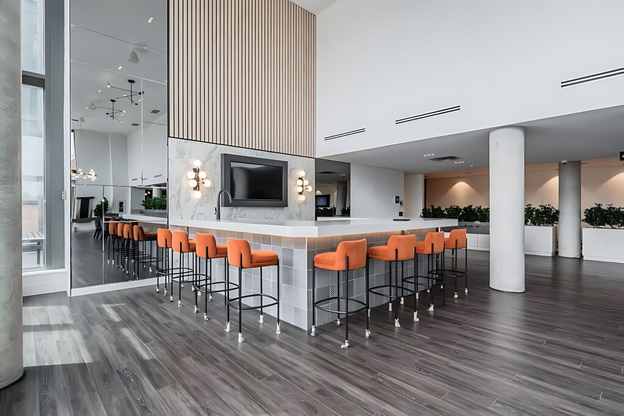 A stylish bar area features a white countertop with a waterfall edge, surrounded by orange bar stools, set against a mirrored and slatted wood accent wall, with modern light fixtures.
