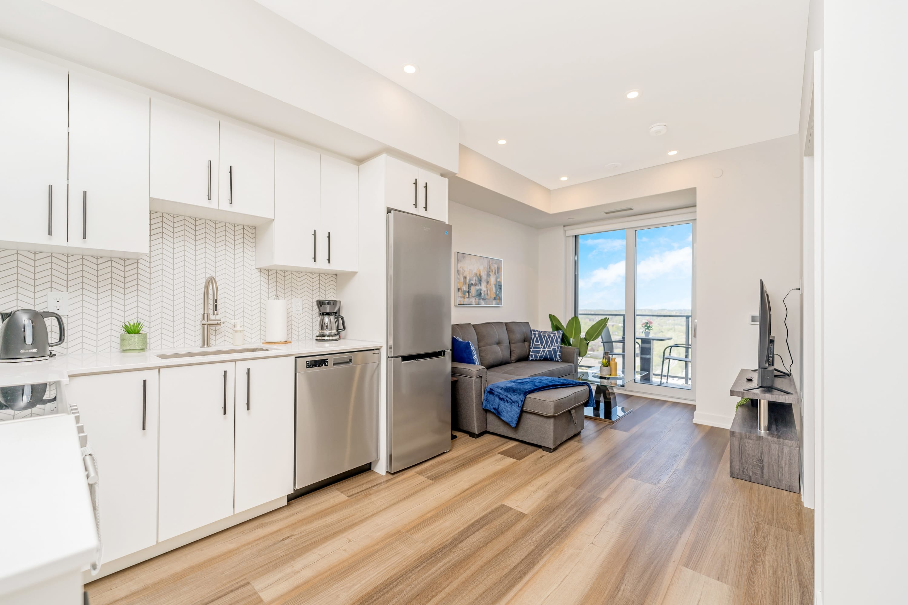 Open-concept view of the kitchen and living room, featuring white kitchen cabinets, stainless steel appliances, a gray sectional sofa, and a balcony with city views in the background.
