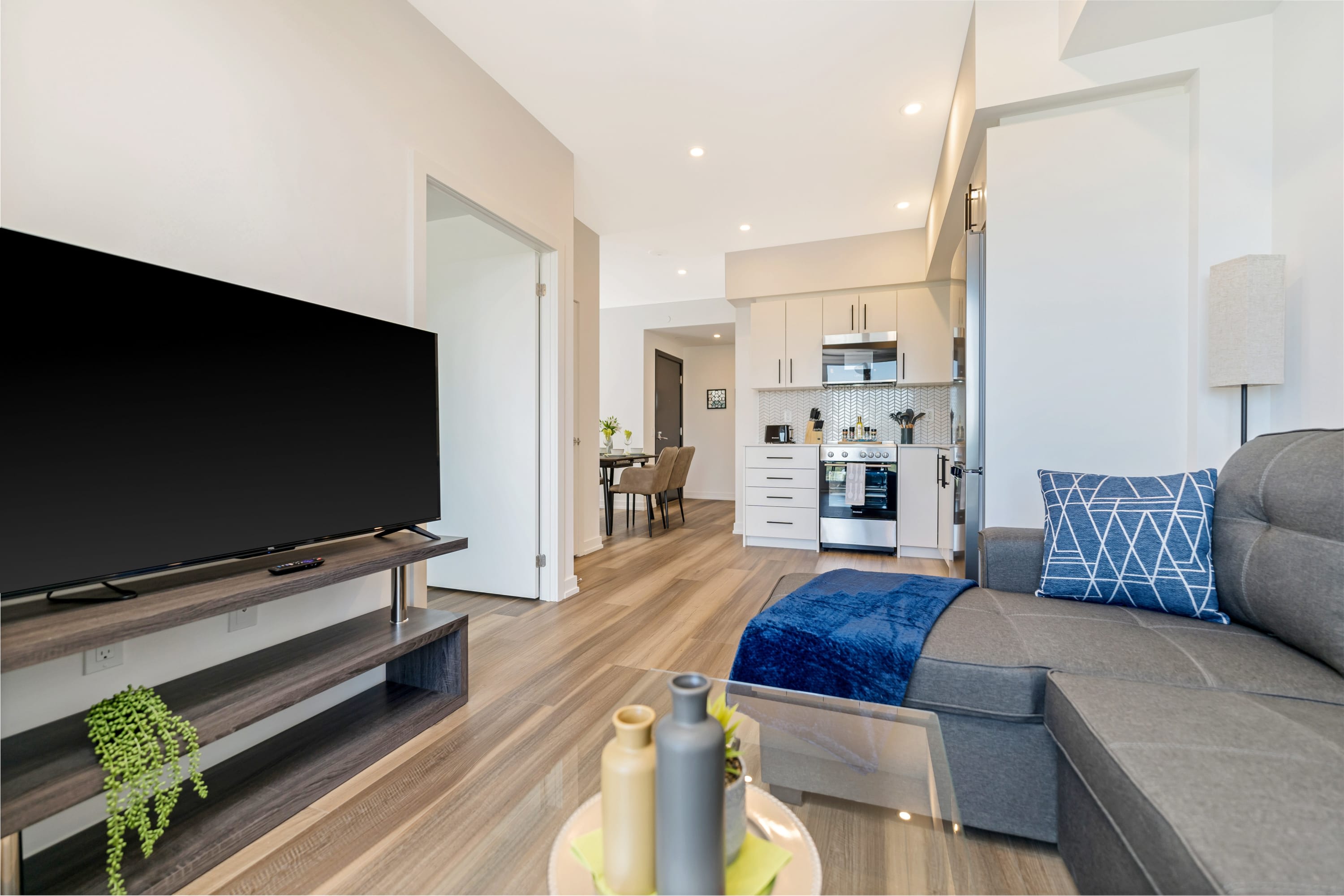 Open-concept living area showing a gray sectional sofa, a large TV on a dark wood stand, and a glimpse of the kitchen and dining area beyond, all with light wood-look flooring.