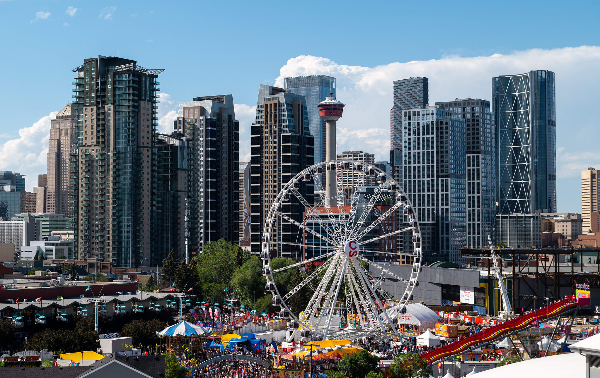 A panoramic city view featuring a prominent white Ferris wheel in the foreground, set against a backdrop of modern high-rise buildings and the iconic Calgary Tower under a blue sky with scattered clouds. The scene captures a lively urban fairground.