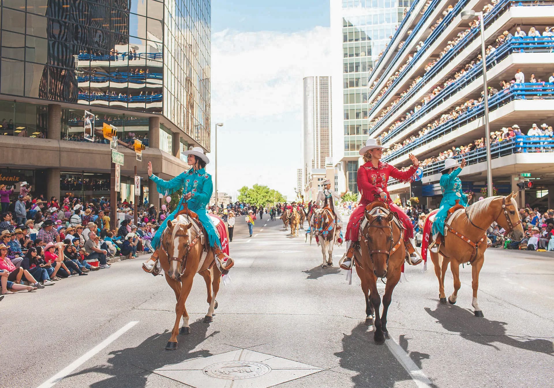 A vibrant city parade scene featuring several people dressed in western attire riding horses down a wide street, flanked by large crowds of spectators and modern high-rise buildings. The atmosphere is festive and lively, capturing a local event.