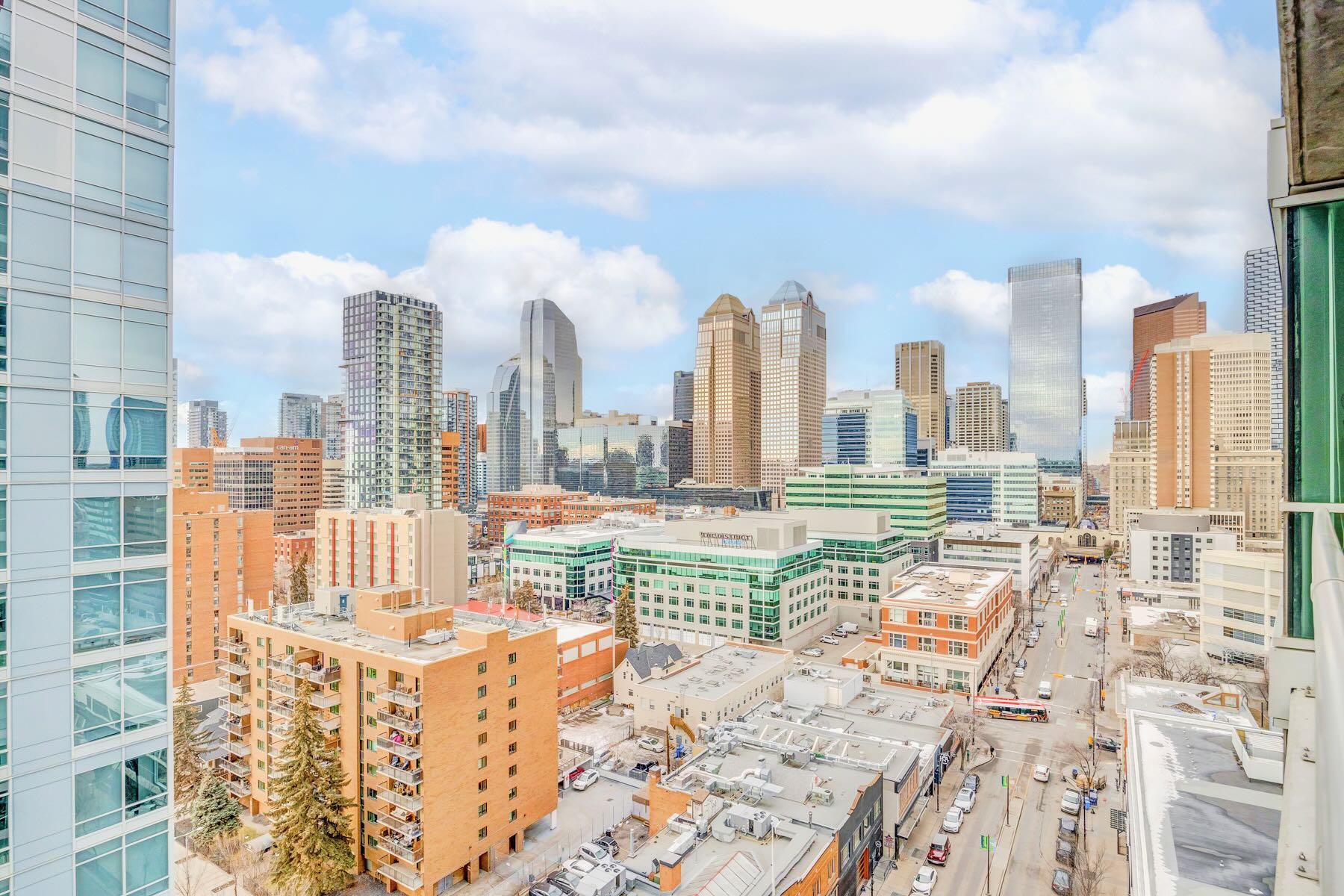 A high-angle city view from a balcony showcasing a dense urban landscape with numerous modern high-rise buildings under a partly cloudy sky. Streets with vehicles are visible below, surrounded by mid-rise residential and commercial structures.