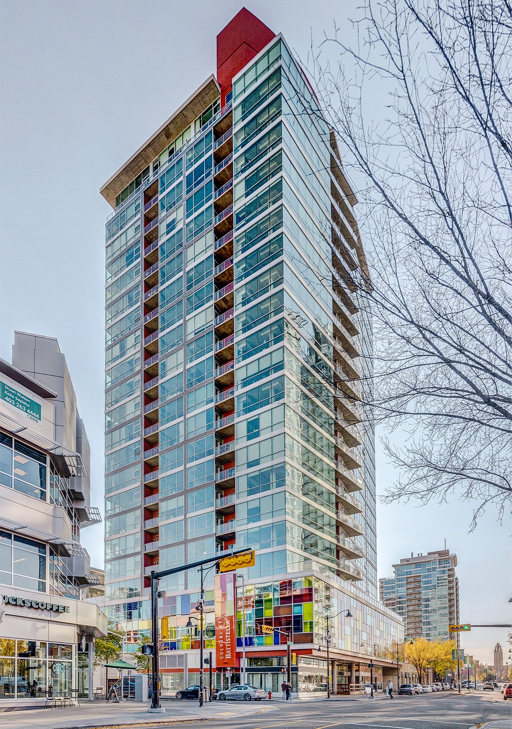 Exterior view of a modern high-rise condominium building with a glass facade and colorful lower-level accents, situated on a city street with other commercial buildings.