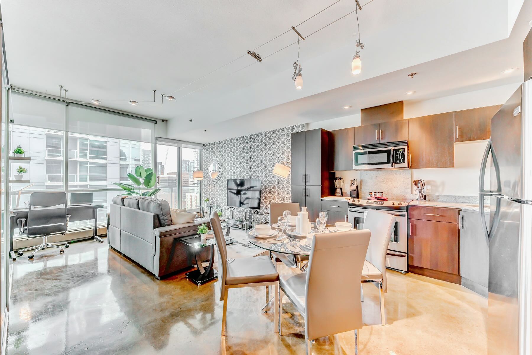A spacious open-plan living area includes a gray sofa, a glass dining table set for four, and a modern kitchen with dark wood cabinetry and stainless steel appliances. Floor-to-ceiling windows provide ample natural light and city views.