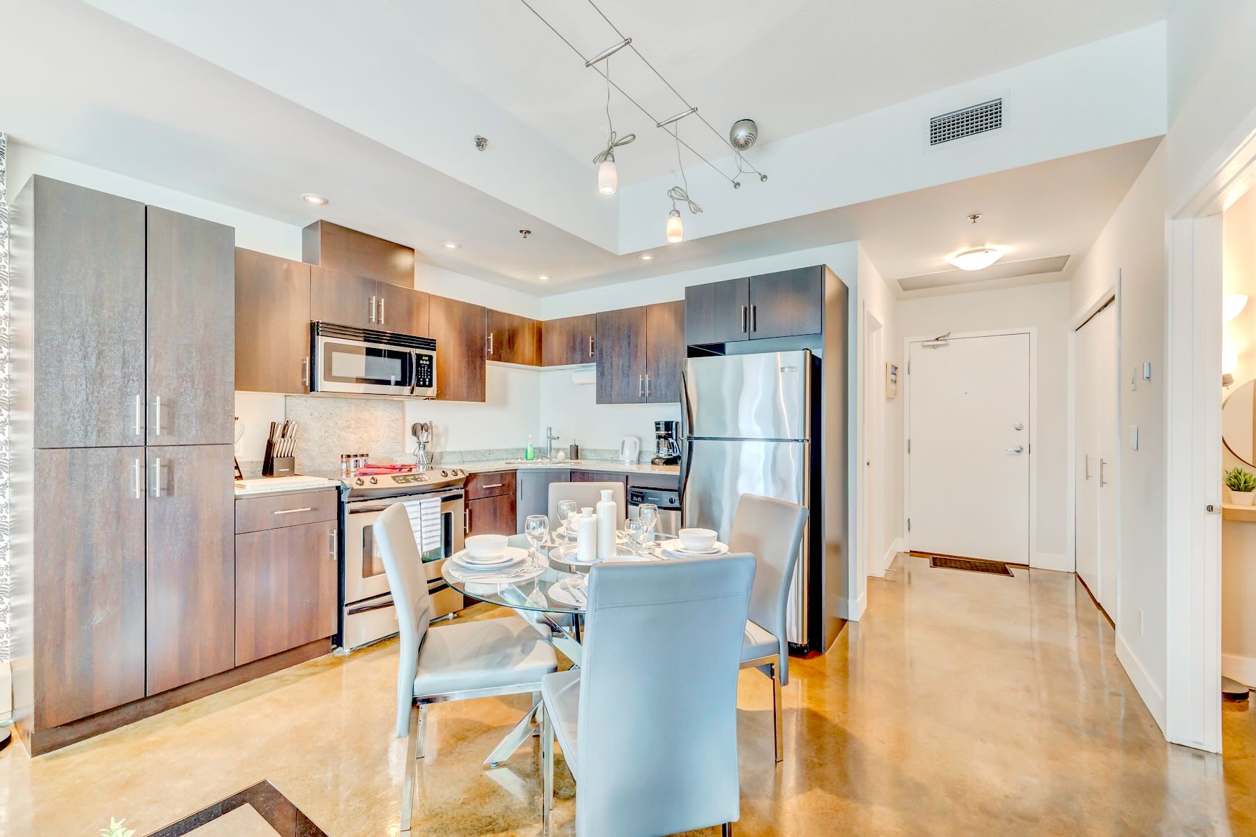 A well-appointed modern kitchen with dark cabinetry and stainless steel appliances, including a microwave and refrigerator, flows into a dining area with a glass table and four chairs. The polished concrete floor extends throughout the open-plan space, leading to the entrance.