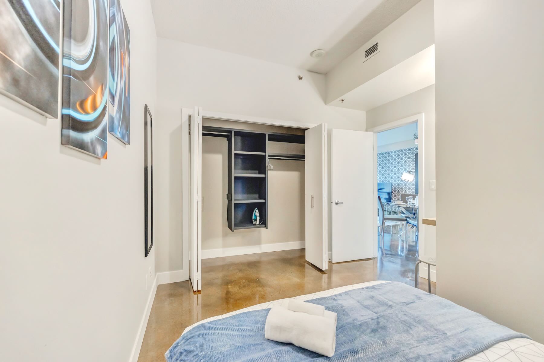 View of the bedroom showcasing a built-in closet with bi-fold doors, featuring shelving and hanging space, next to the blue velvet bed and polished concrete floor.
