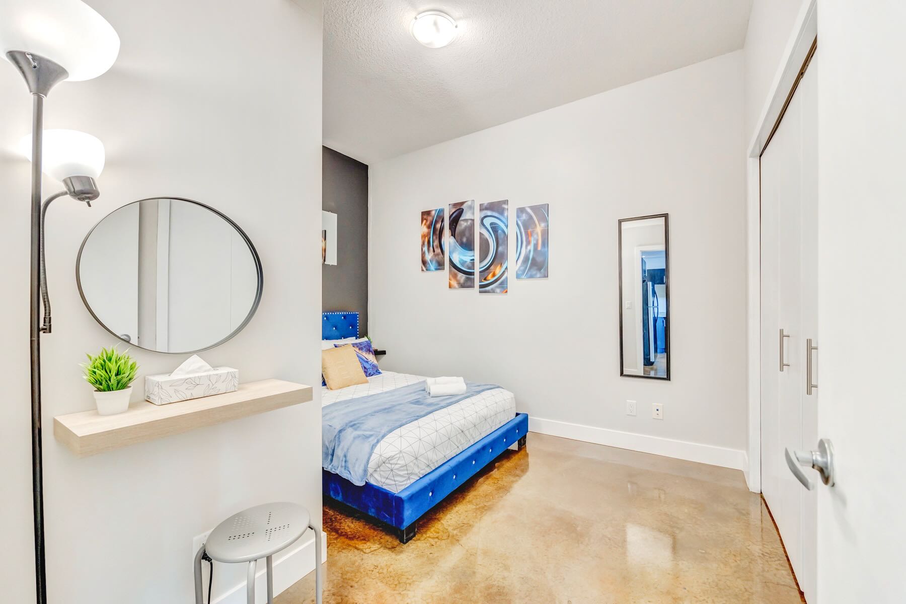 Another angle of the bedroom, highlighting a full-length mirror, abstract wall art, and a small vanity area with a round mirror and stool, all on a polished concrete floor.