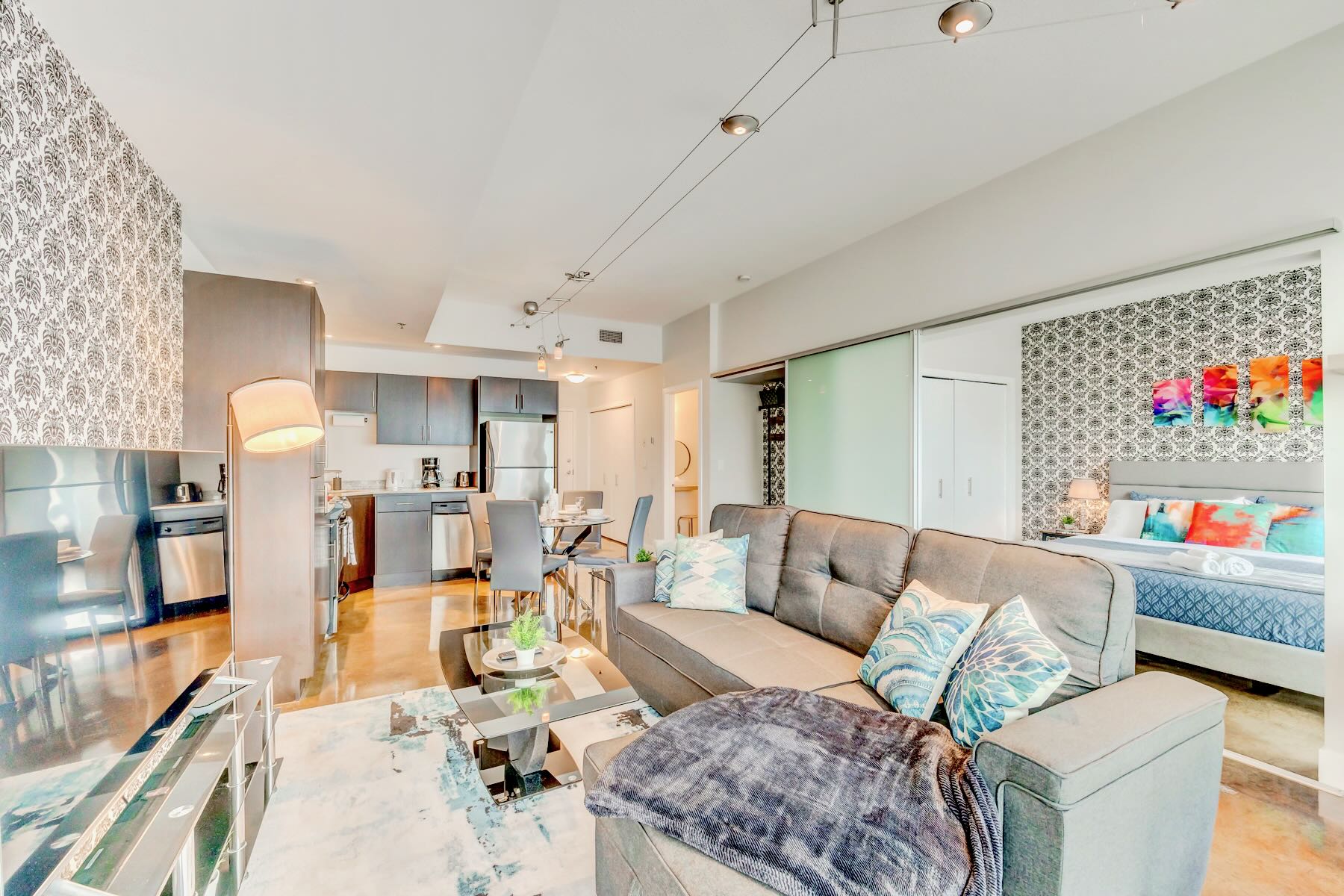 An open-concept living space features a gray sectional sofa, a glass coffee table, and a large TV, with a modern kitchen and dining area visible in the background. A bedroom with patterned wallpaper is partially visible through sliding glass doors.