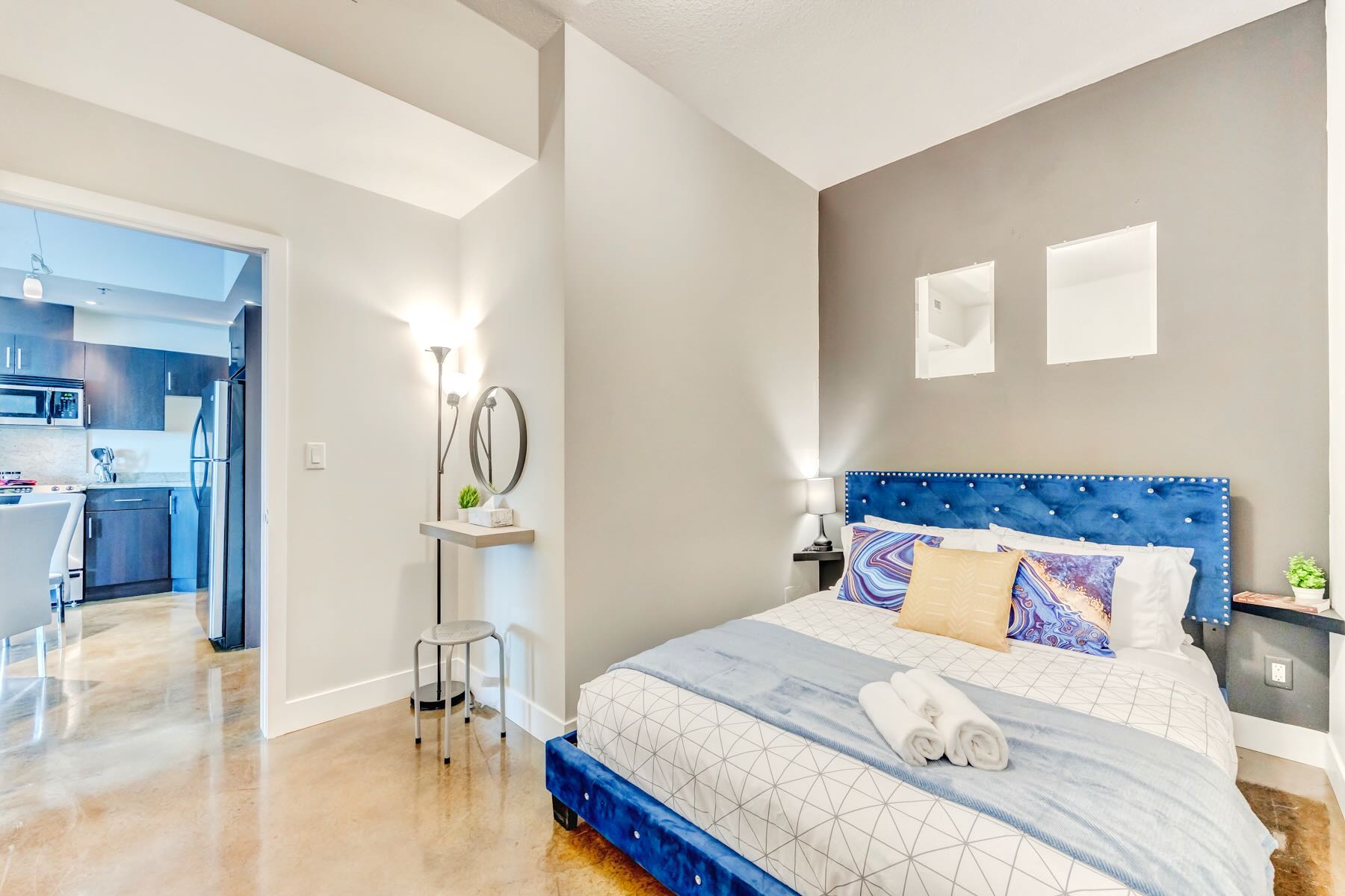 Stylish bedroom featuring a blue velvet bed with a geometric pattern duvet, a dark gray accent wall, and a polished concrete floor, with a glimpse of the kitchen area through the doorway.