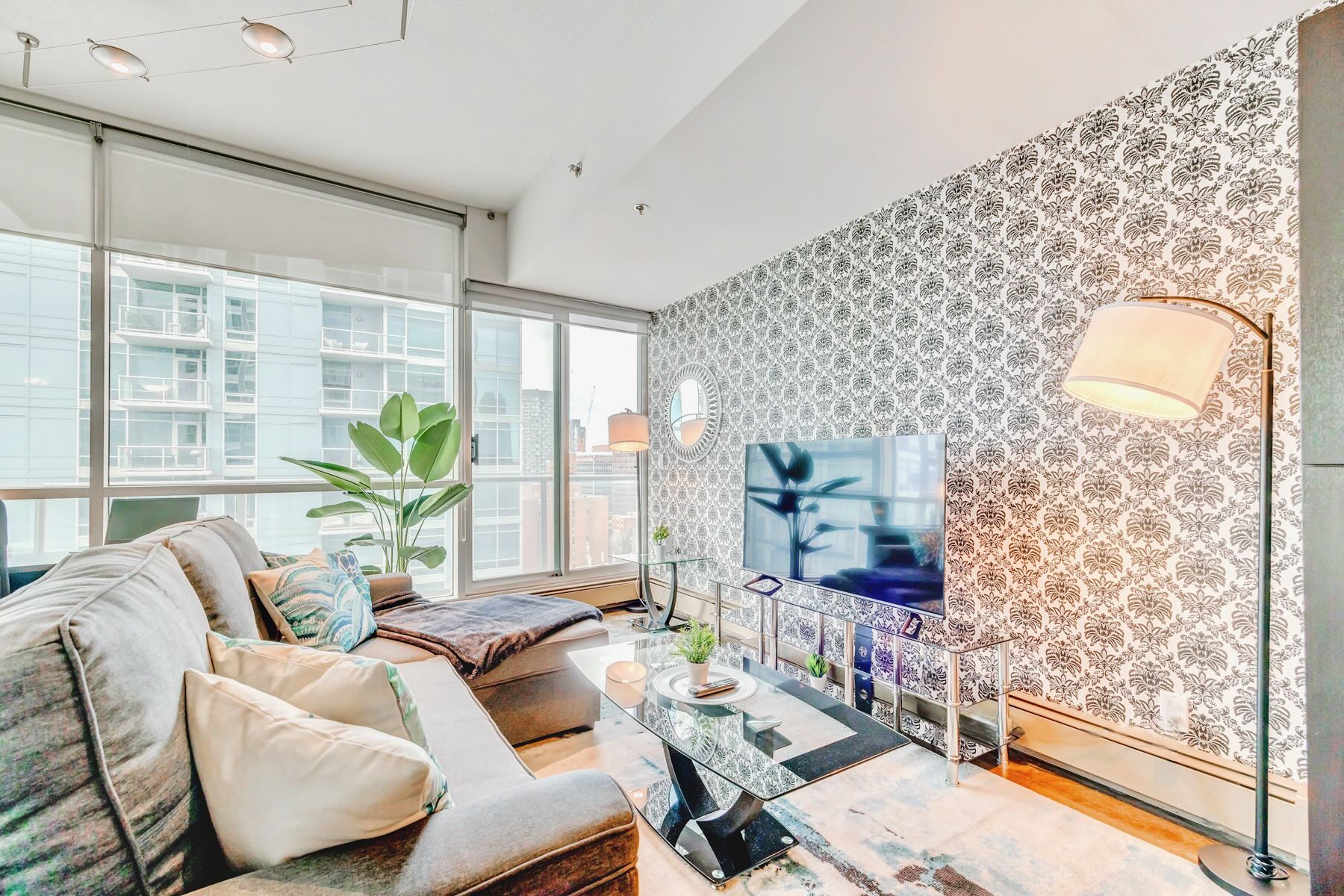 Cozy living room corner with a gray sectional sofa, a large wall-mounted TV against a black and white patterned wallpaper, and expansive windows offering city views. A tall floor lamp provides ambient lighting.
