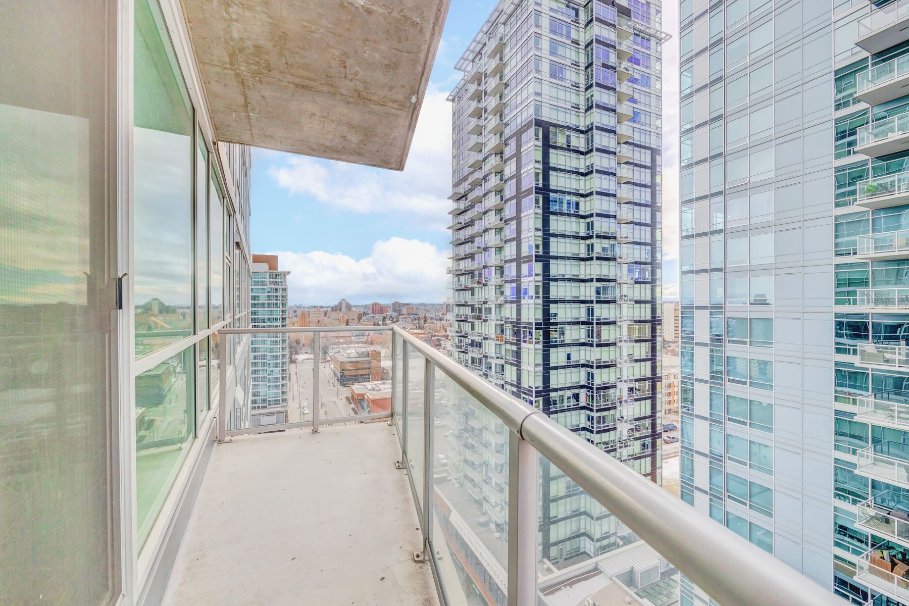 Outdoor balcony with a clear glass railing, offering a high-rise view of modern city buildings and a partly cloudy sky.