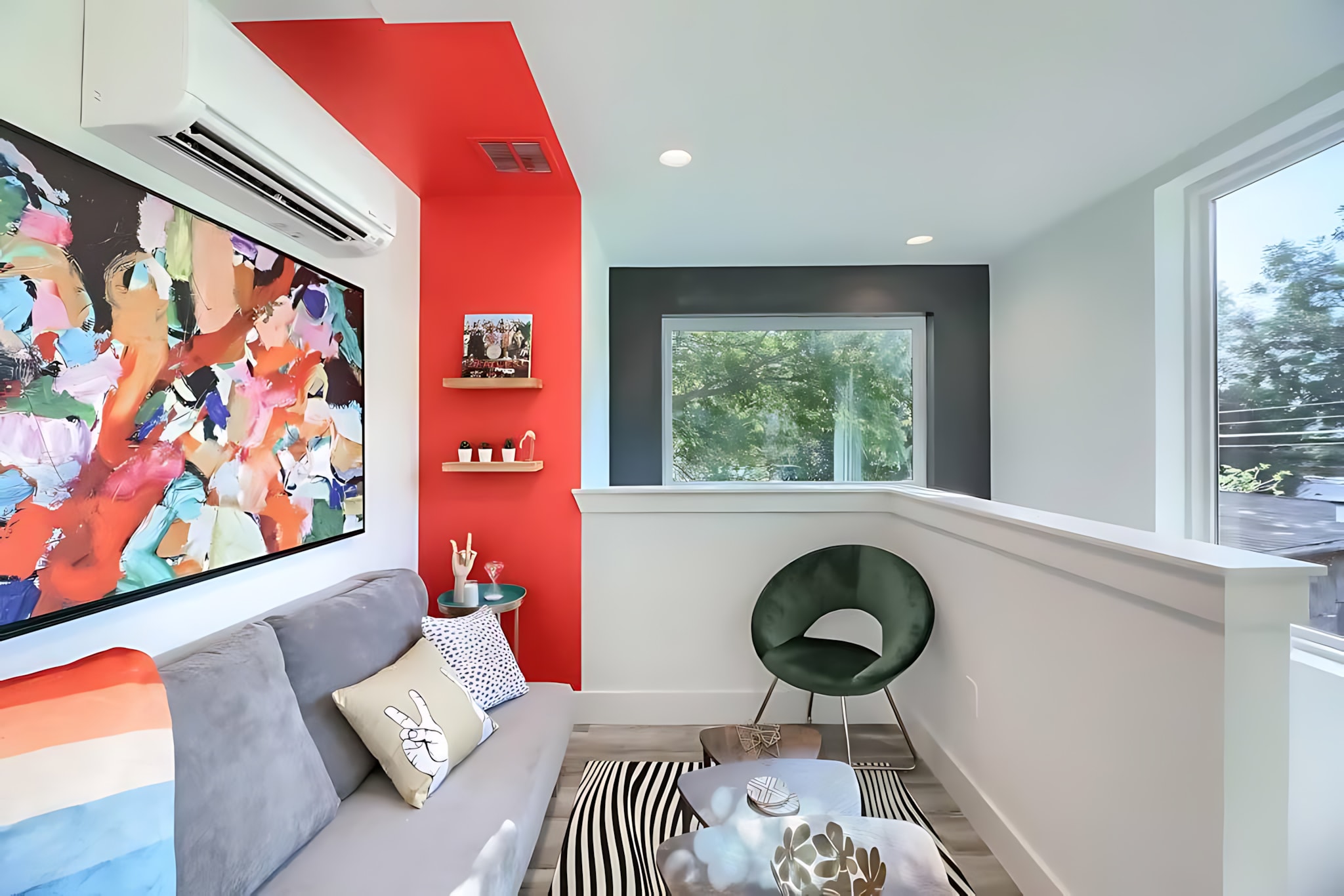 Vibrant upstairs lounge area with a grey sofa, colorful abstract artwork, and a striking red accent wall. A black and white striped rug defines the seating space, which includes a small green chair and offers natural light from a window.