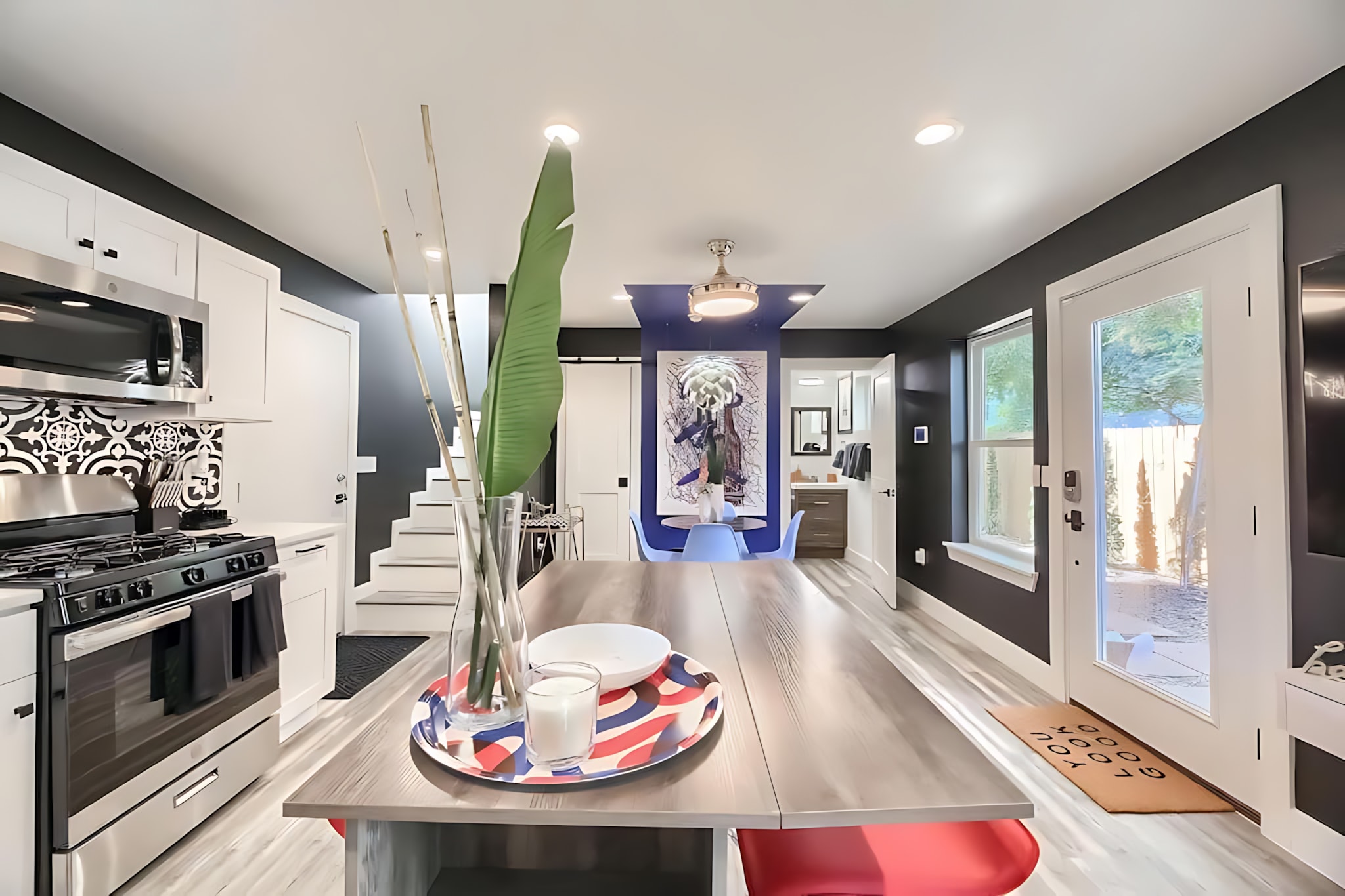 Open-concept living space showcasing a modern kitchen island with red stools, leading to a dining area with a black marble table and blue chairs. The vibrant blue accent wall with abstract art adds a pop of color, and a staircase is visible in the background.