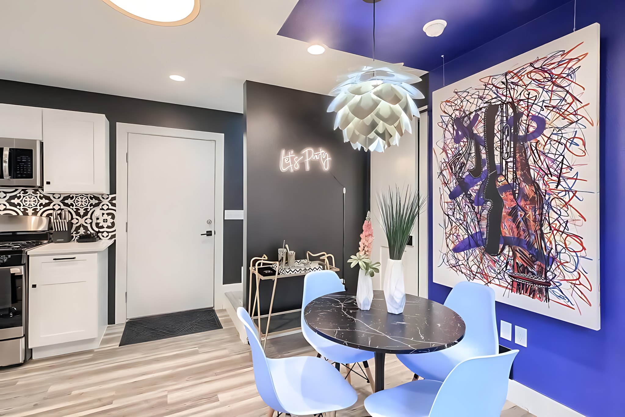 Eclectic dining area featuring a black marble-top table and light blue chairs, positioned under a distinctive white pendant light. A bold blue accent wall with abstract art contrasts with the dark walls and white kitchen cabinets in the background.