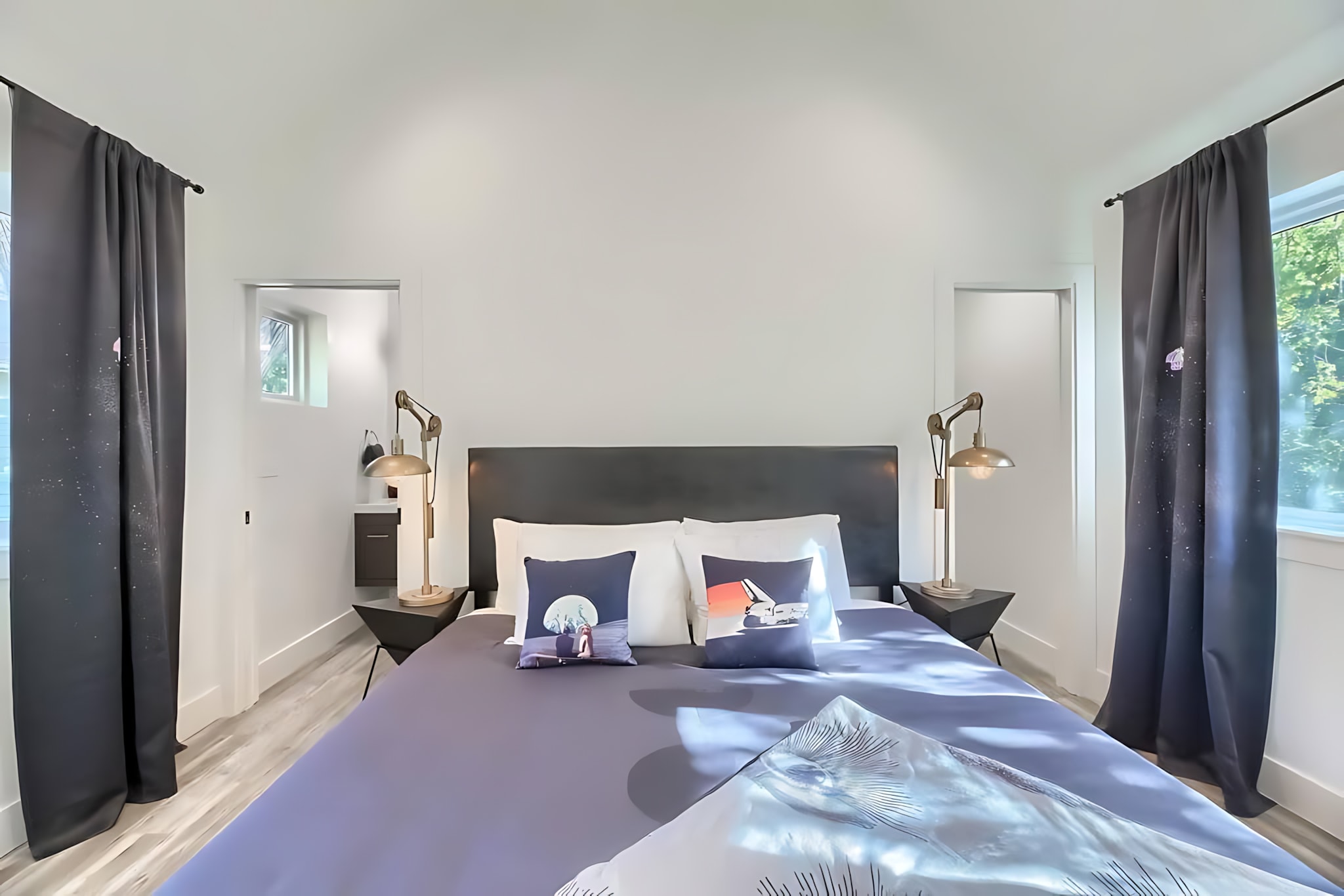 A modern bedroom with a large bed featuring a dark headboard, two nightstands with industrial-style lamps, and dark gray curtains framing windows on either side.
