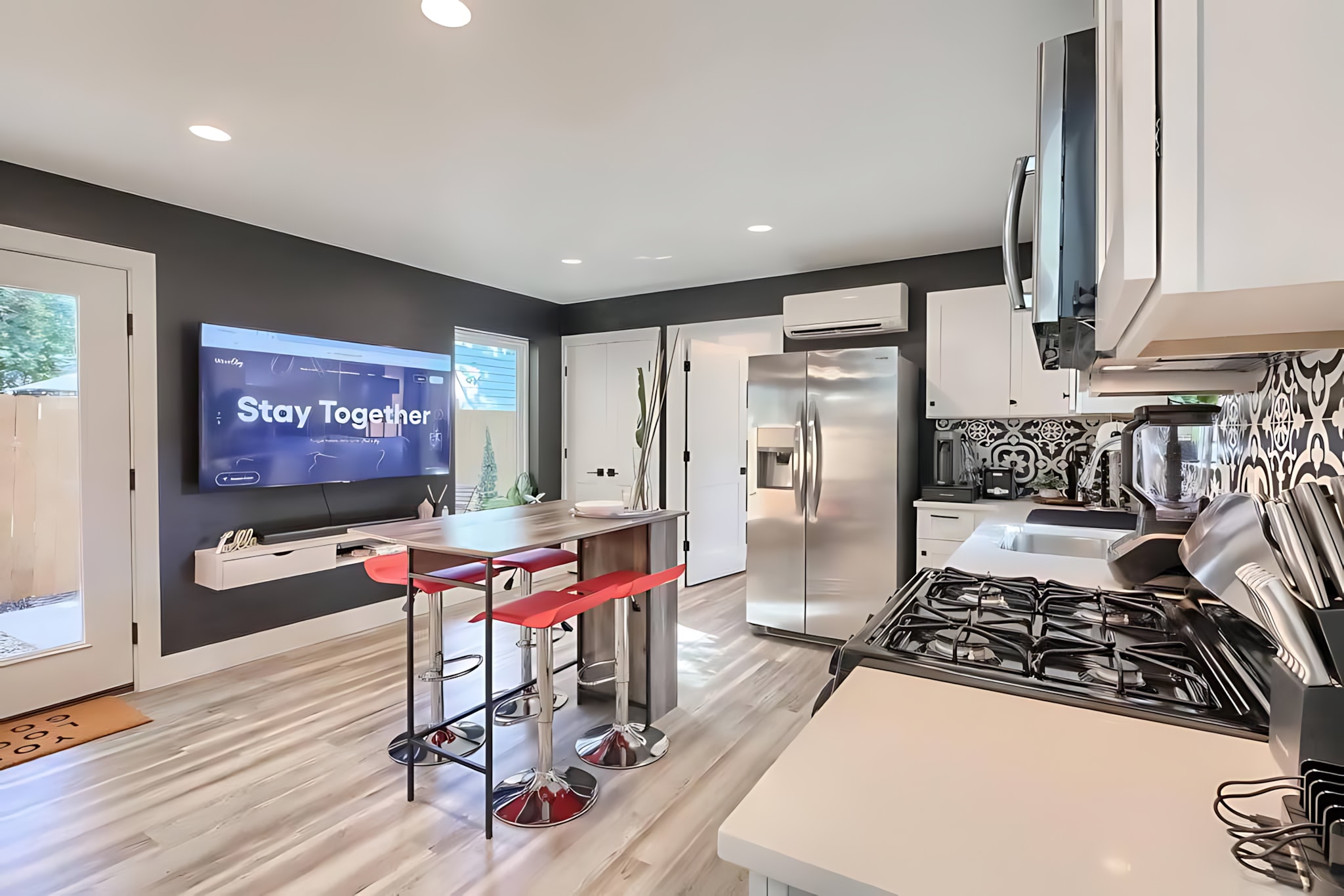 Modern kitchen with white cabinets, stainless steel appliances, and a large island with red bar stools. A wall-mounted TV is visible on the dark accent wall, and a glass door leads to the outside.