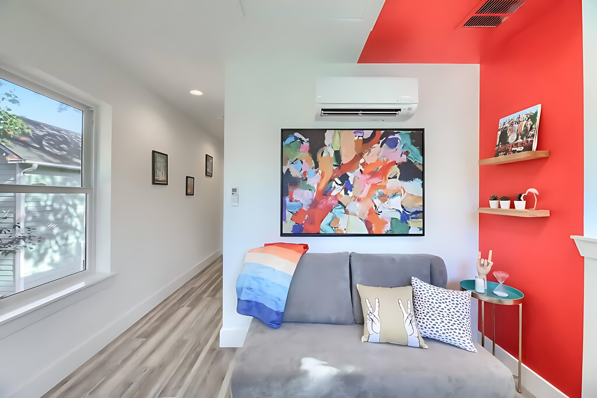 A vibrant living space featuring a gray chaise lounge sofa with colorful pillows and a blanket, set against a white wall with a large abstract painting, and a bold red accent wall with floating shelves.