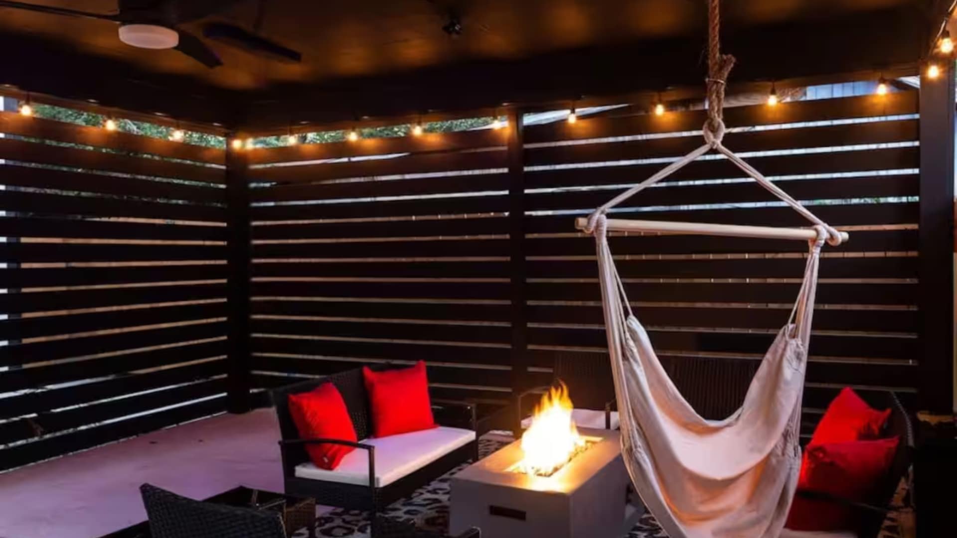Close-up of a covered outdoor patio featuring a central fire pit, surrounded by black wicker seating with bright red cushions. A comfortable hanging chair and string lights add to the inviting ambiance.