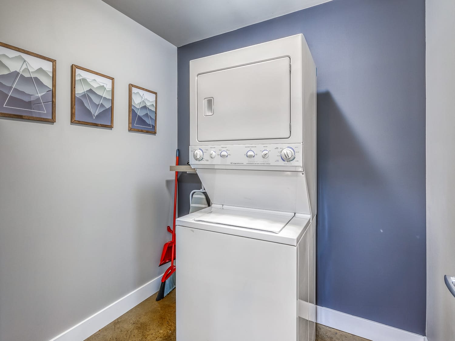 Compact laundry area featuring a white stacked washer and dryer unit. The walls are painted in a two-tone scheme of gray and blue, with framed abstract artworks on the gray wall.