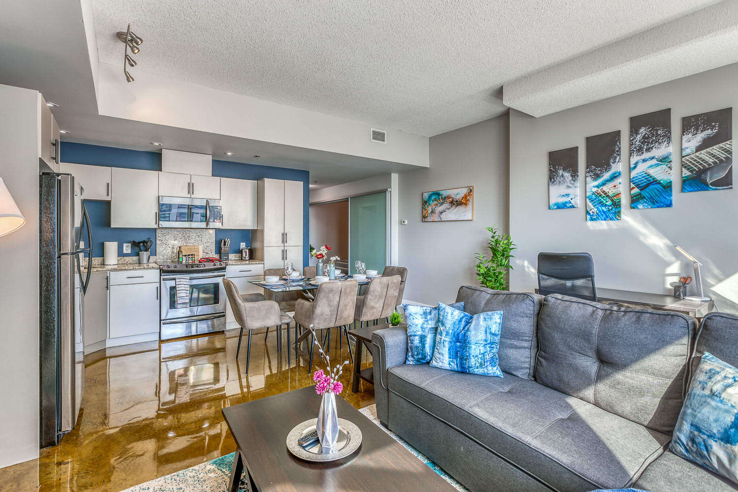 Open-concept living area with a gray sofa, a dark coffee table, and a blue and gray abstract rug. The space flows into a dining area and a kitchen with white cabinets and stainless steel appliances.