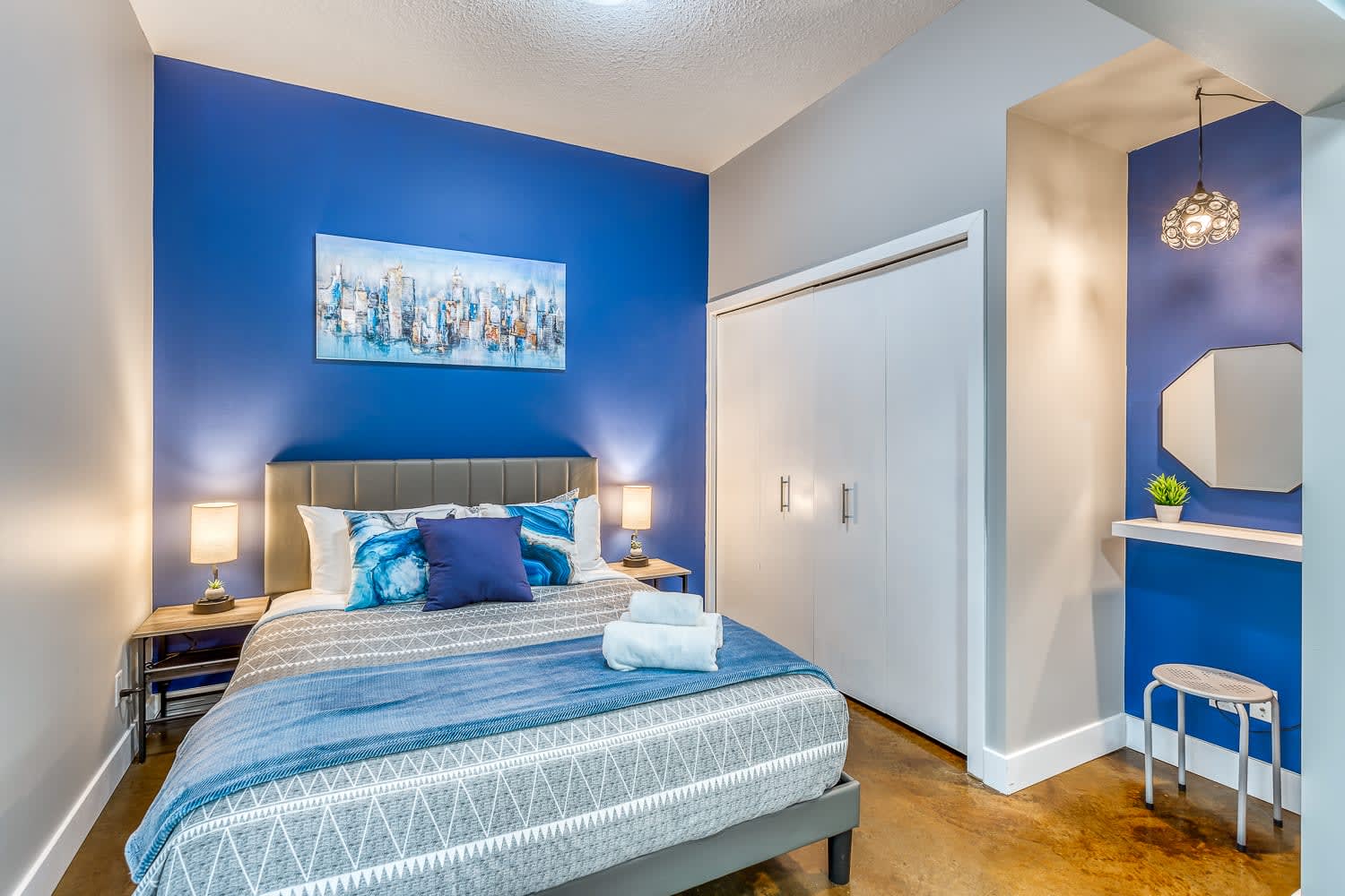 Vibrant bedroom with a queen-sized bed featuring blue and gray bedding, flanked by two nightstands with lamps. A large blue accent wall is adorned with a cityscape painting, and a white closet is visible.