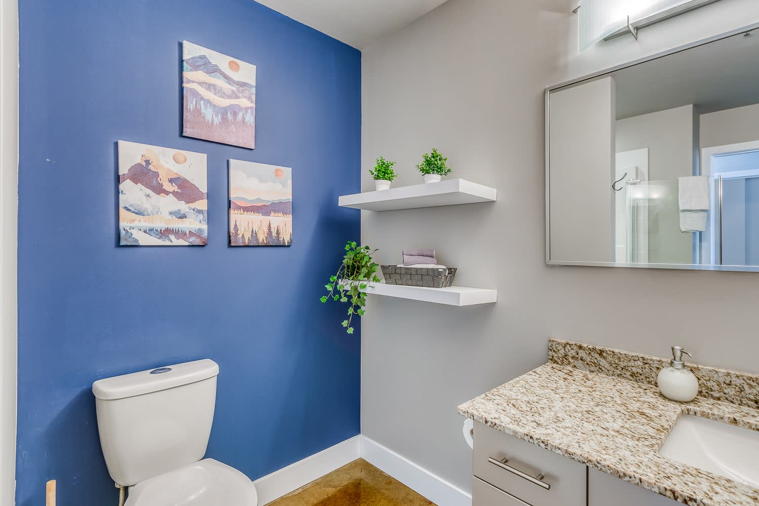 Corner of a modern bathroom featuring a white toilet against a vibrant blue accent wall, adorned with three framed abstract mountain artworks. A light-colored granite vanity and mirror are partially visible on the adjacent gray wall.