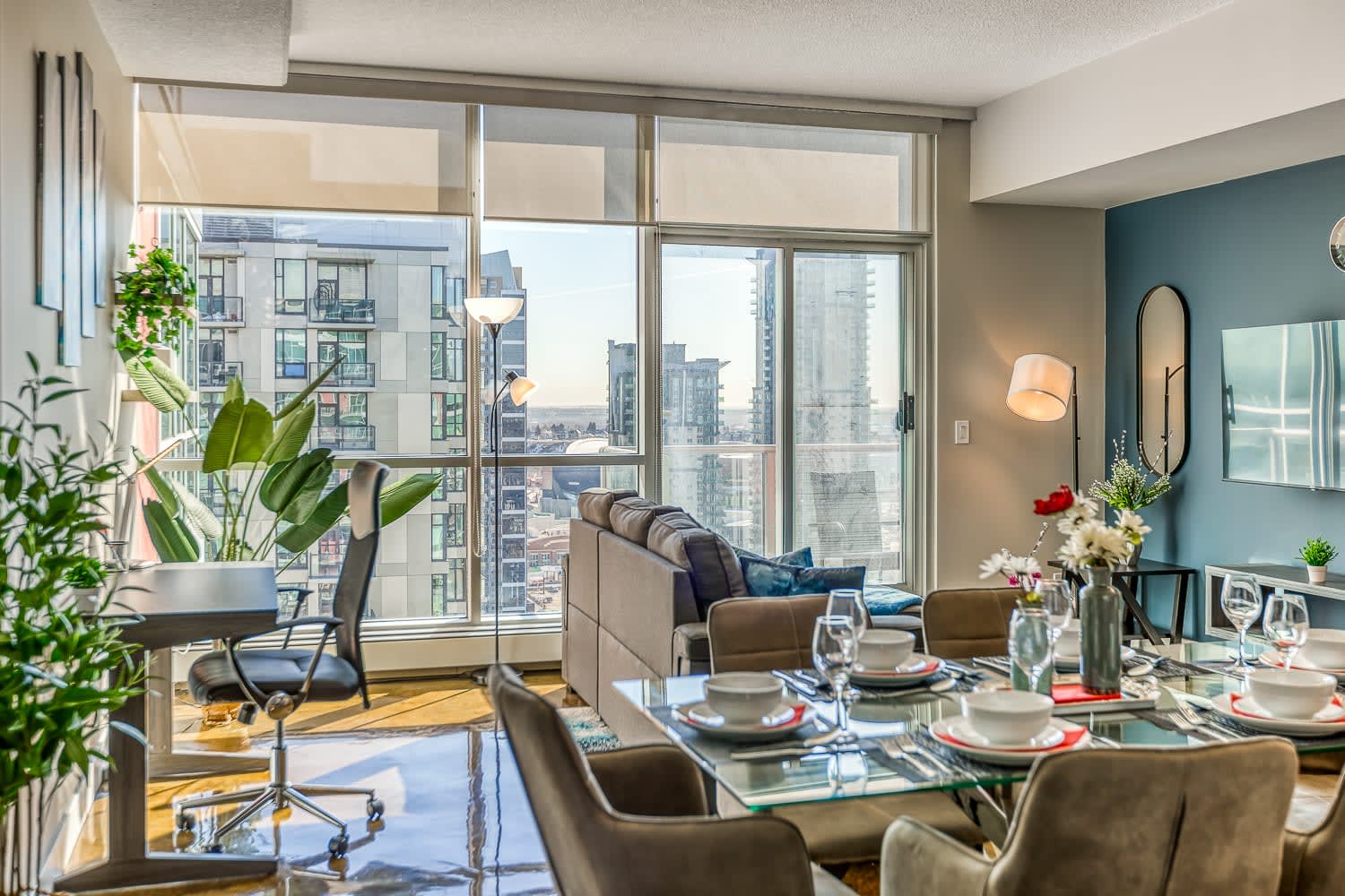 Bright living and dining area with a gray sofa and a glass dining table set for six. Large windows provide abundant natural light and city views, while a dedicated workspace with a desk and chair is visible on the left.