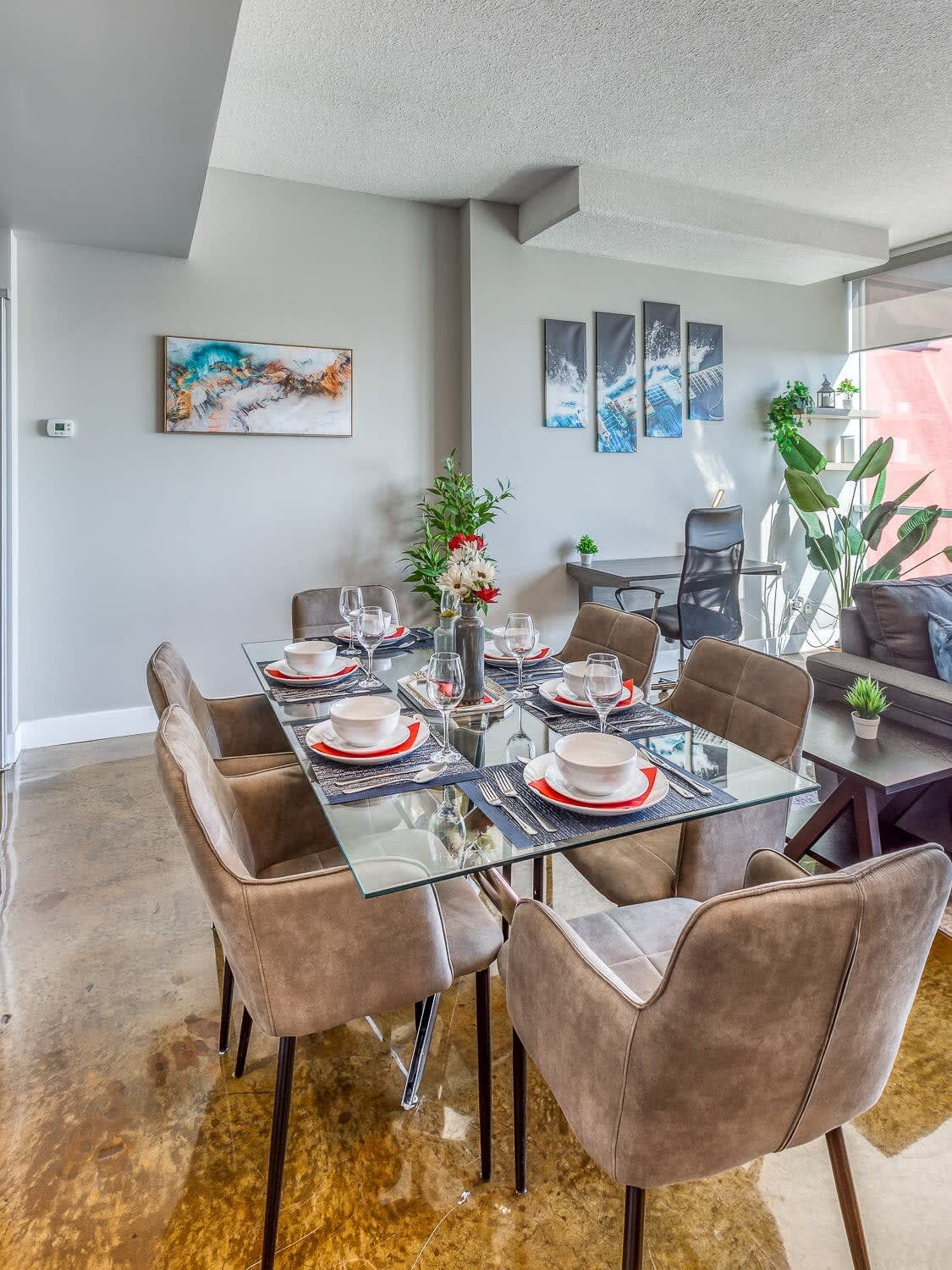 Elegant dining area featuring a glass-top table with six gray upholstered chairs, set with modern place settings. The space is adorned with contemporary wall art and a small floral centerpiece.