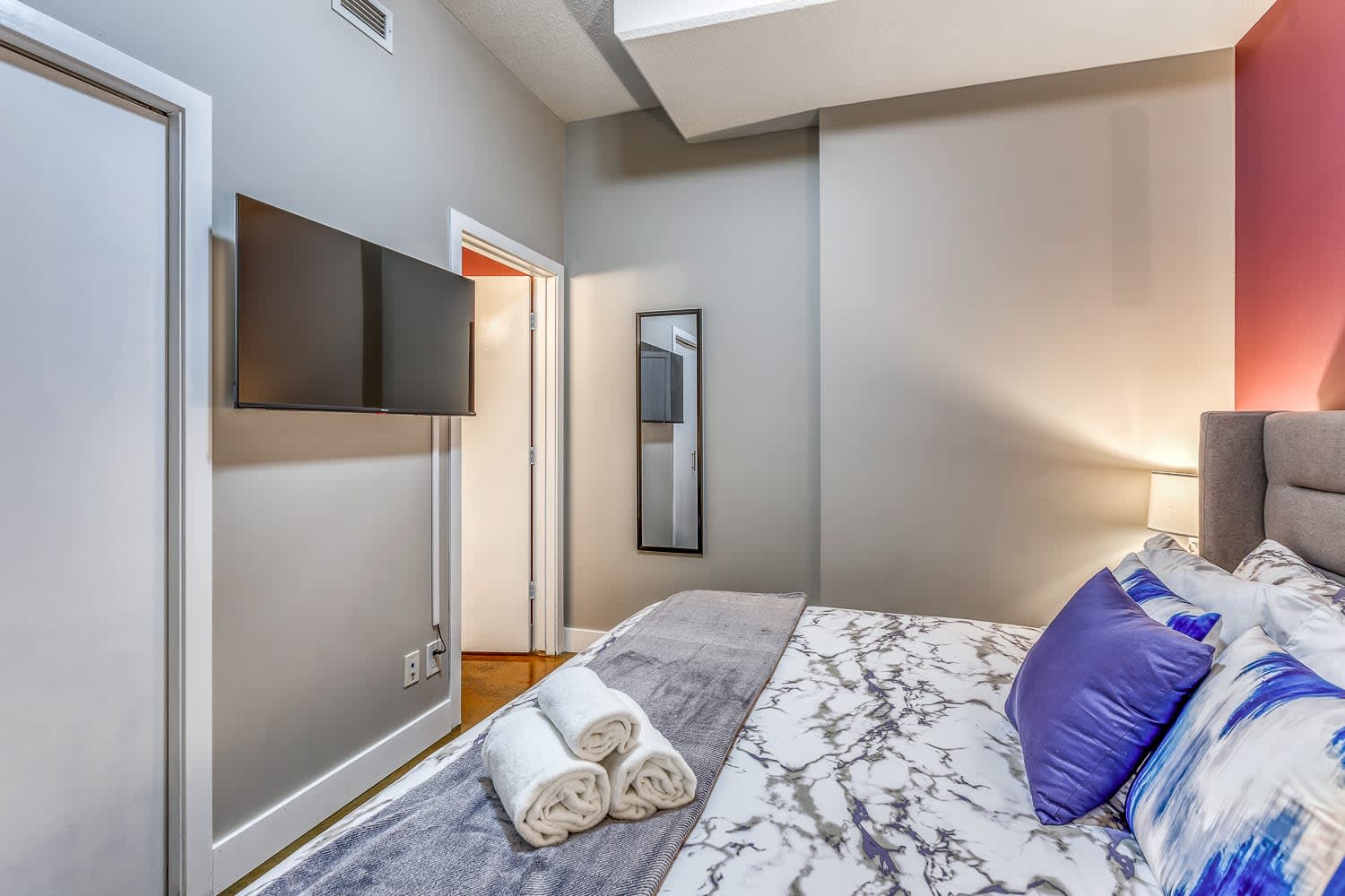 View of bedroom 1 showing the gray upholstered bed with marble-patterned bedding and a wall-mounted flat-screen TV. A full-length mirror is visible through the open doorway, reflecting the room's light gray walls.