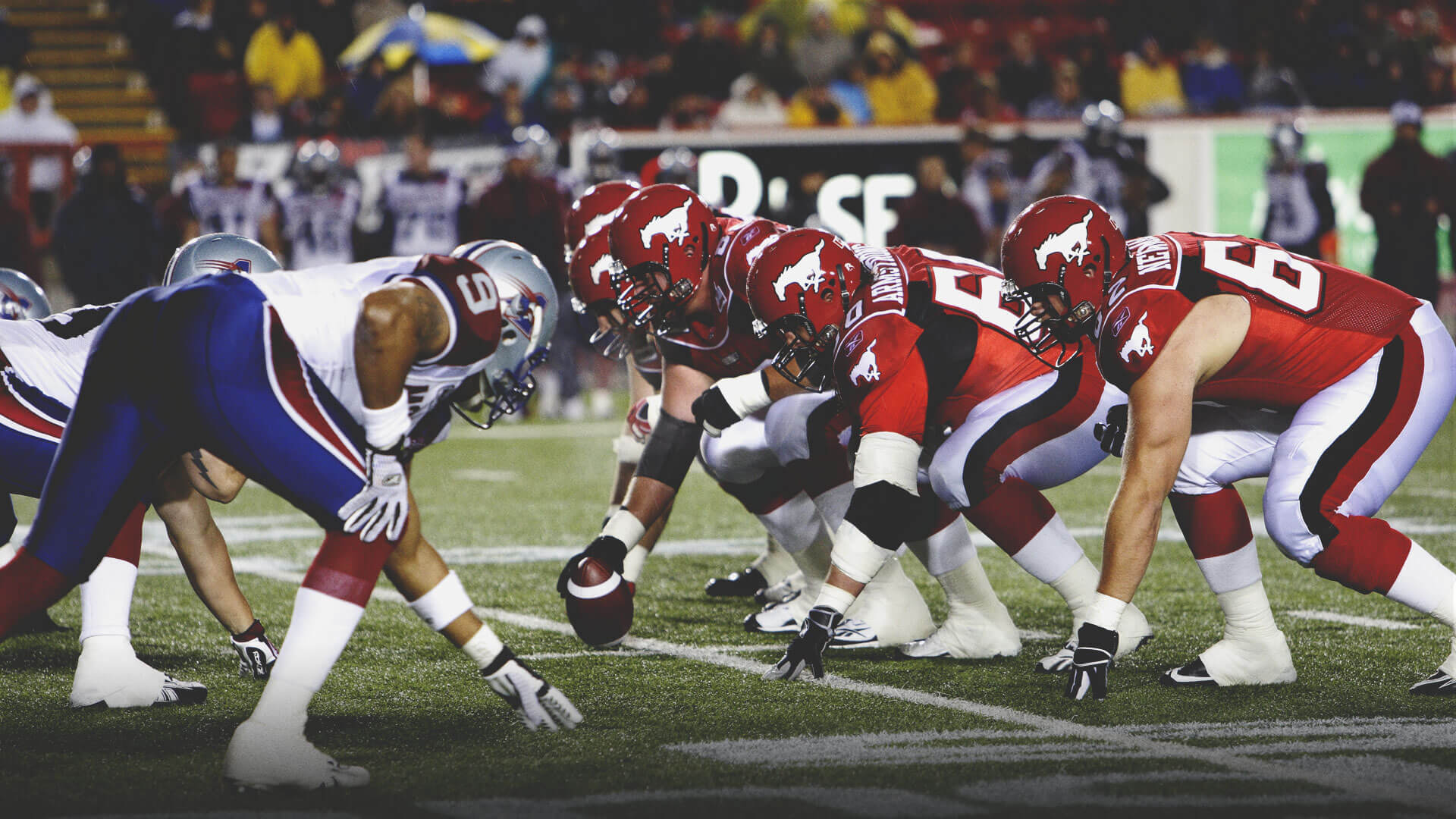 Dynamic shot of a football game in progress on a green turf field, with players in red and white uniforms lined up against opponents in blue and white. Spectators are visible in the background under stadium lights.