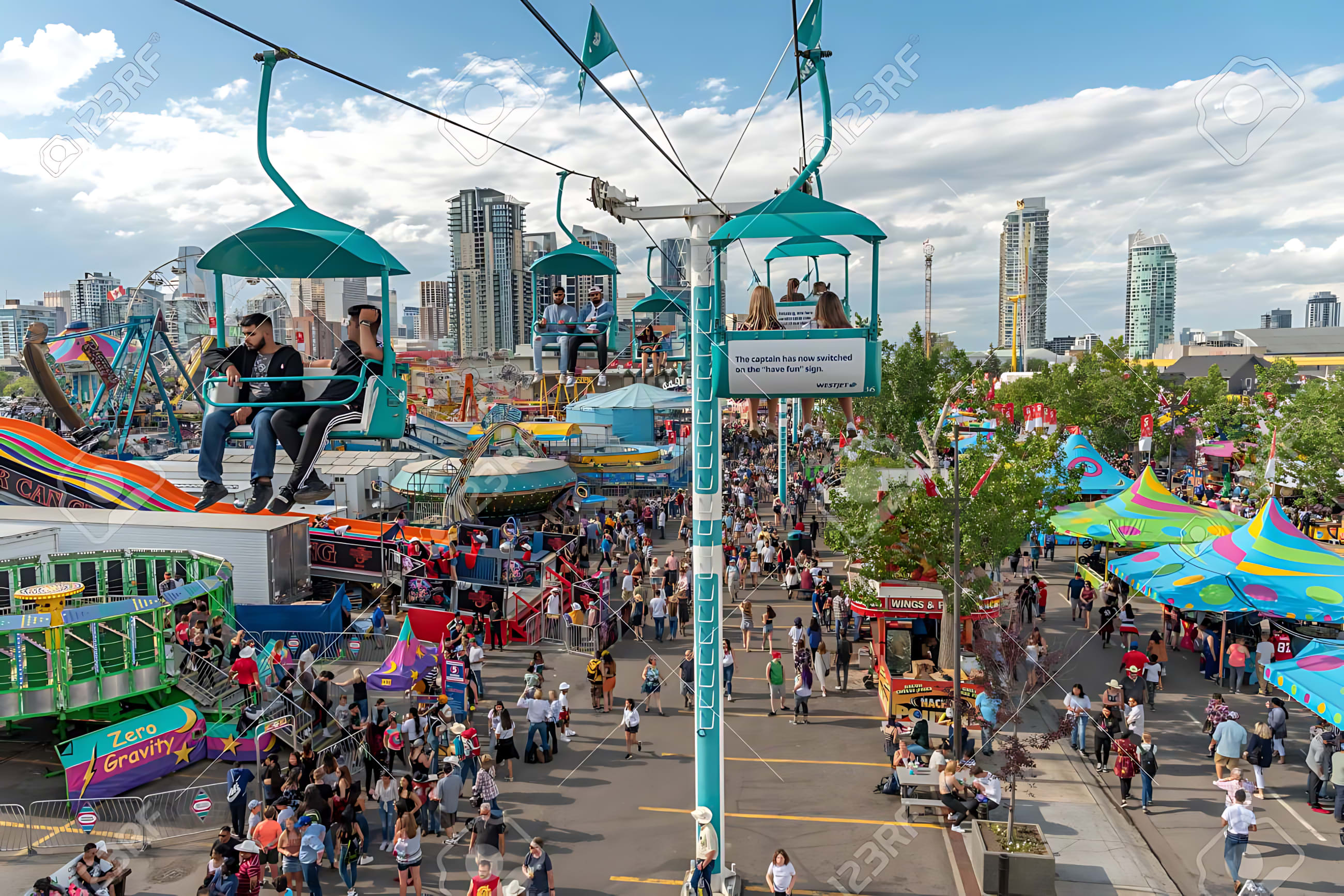 Panoramic view of a bustling amusement park with a chairlift carrying people overhead, surrounded by various rides and food stalls. A city skyline is visible in the background under a partly cloudy sky.