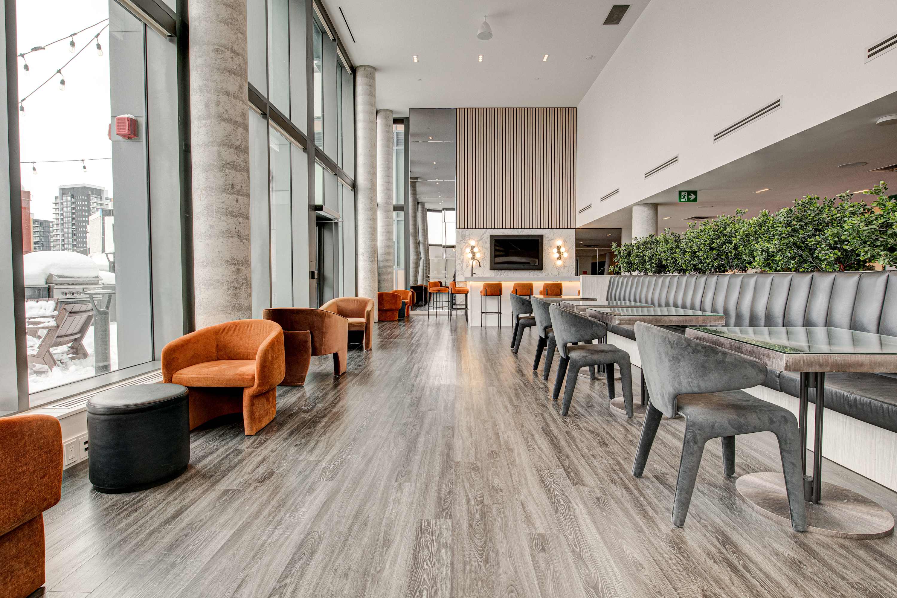 A spacious communal dining and lounge area features a long banquette with grey seating, square tables with dark chairs, and several orange armchairs by large windows, all set on light wood-look flooring.
