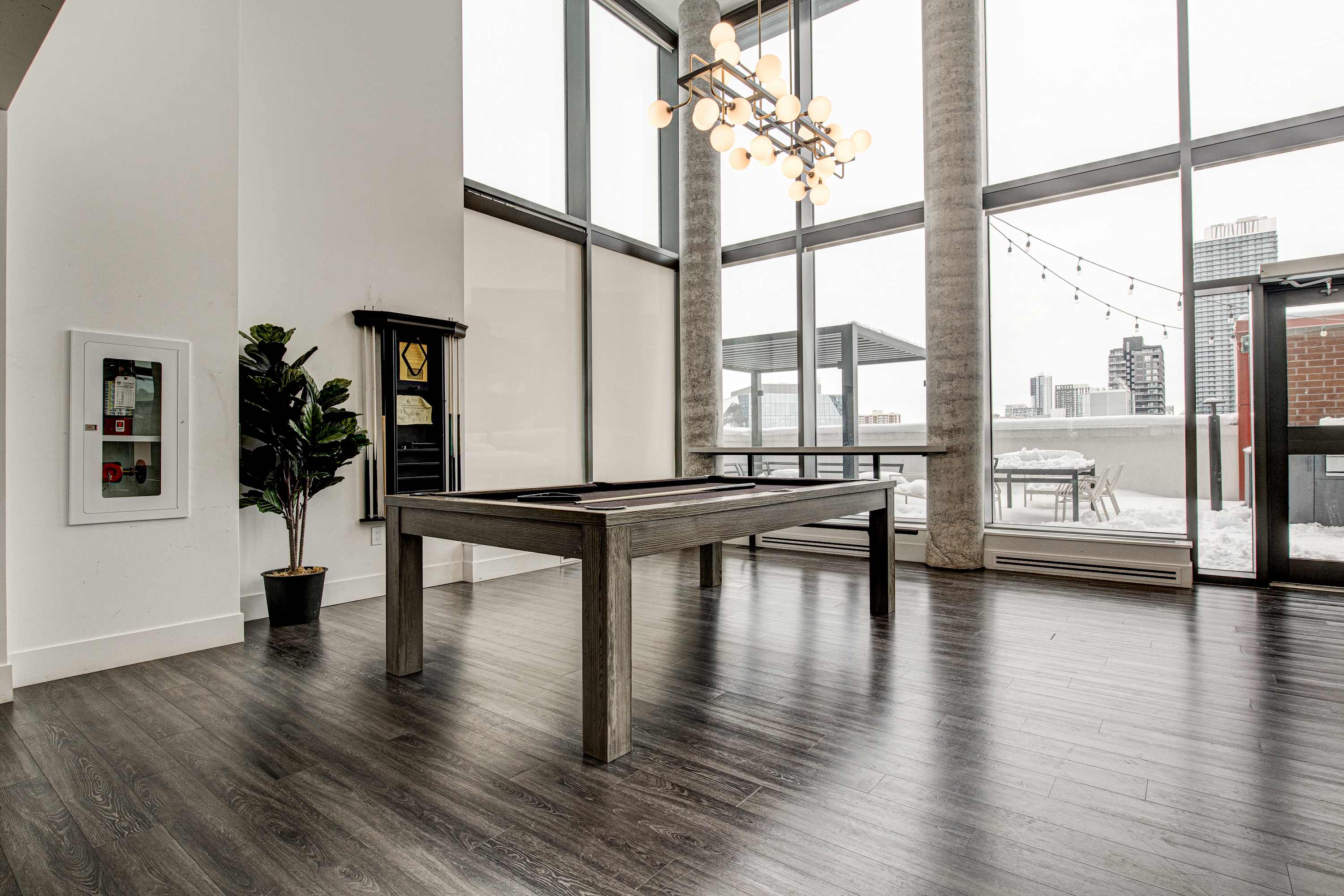 A contemporary game room features a dark wood pool table at its center, with dark wood flooring, tall concrete pillars, and large windows offering views of a snowy outdoor patio. A modern chandelier hangs overhead.