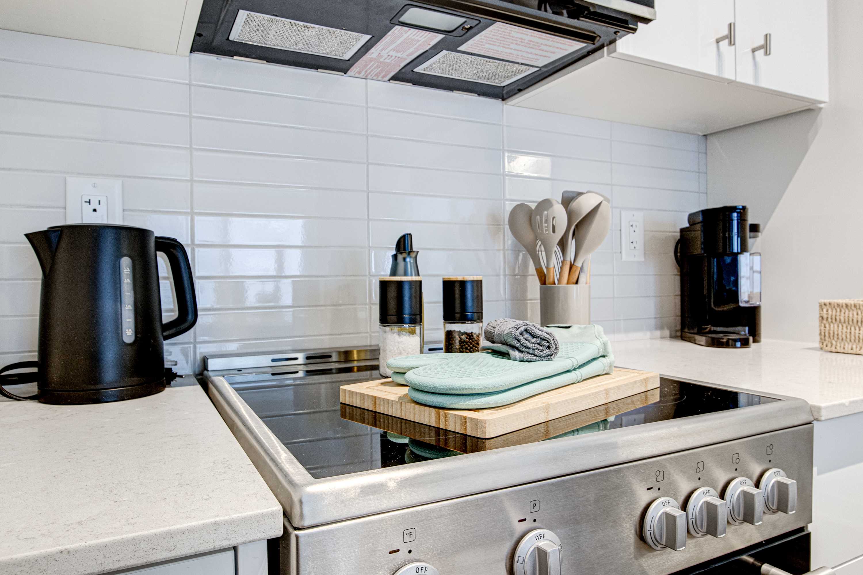 Contemporary kitchen with a stainless steel electric stove, white subway tile backsplash, and light-colored countertops, featuring a black kettle, coffee maker, and cooking utensils.