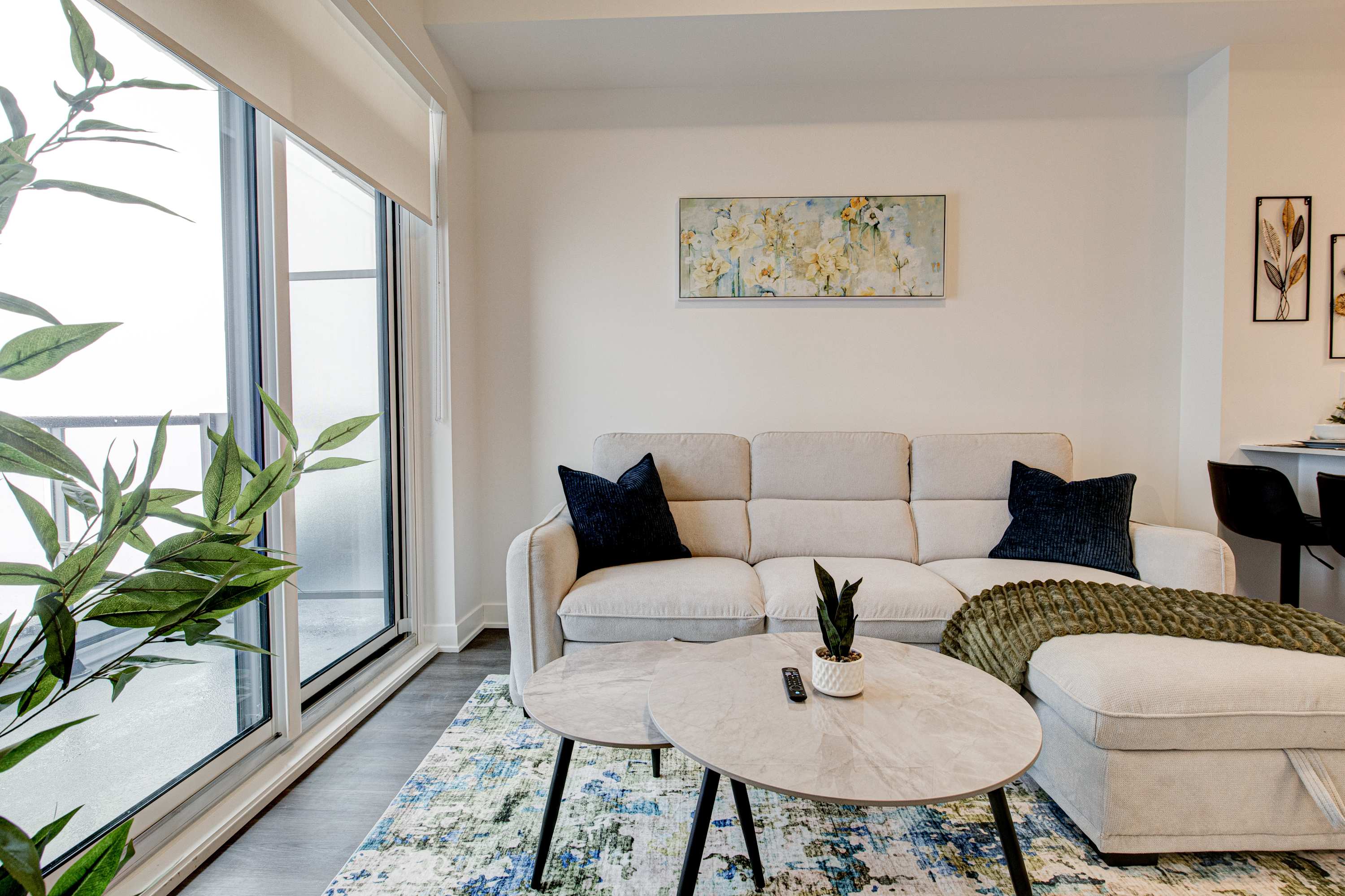 A bright living room features a light beige sectional sofa with dark blue throw pillows and a green blanket, positioned on a colorful abstract rug. Large windows overlook a city view, and a round marble coffee table sits in front of the sofa.