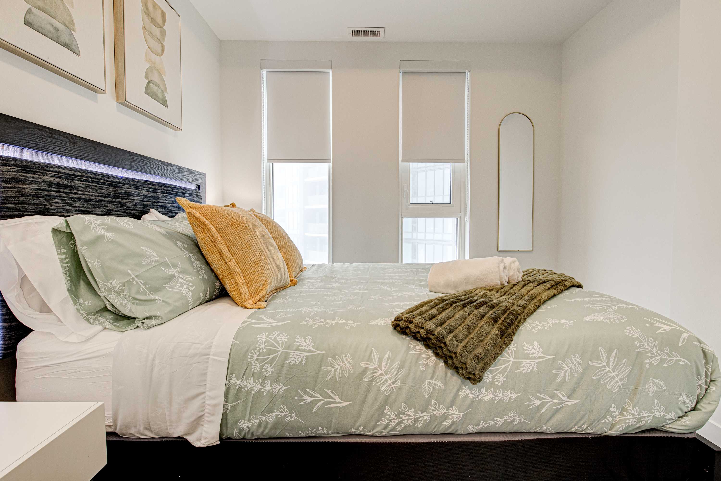 View of the bedroom from the side, highlighting the light green floral duvet cover, yellow throw pillows, and a dark green textured blanket on a dark wood bed frame, with two tall windows on the right.