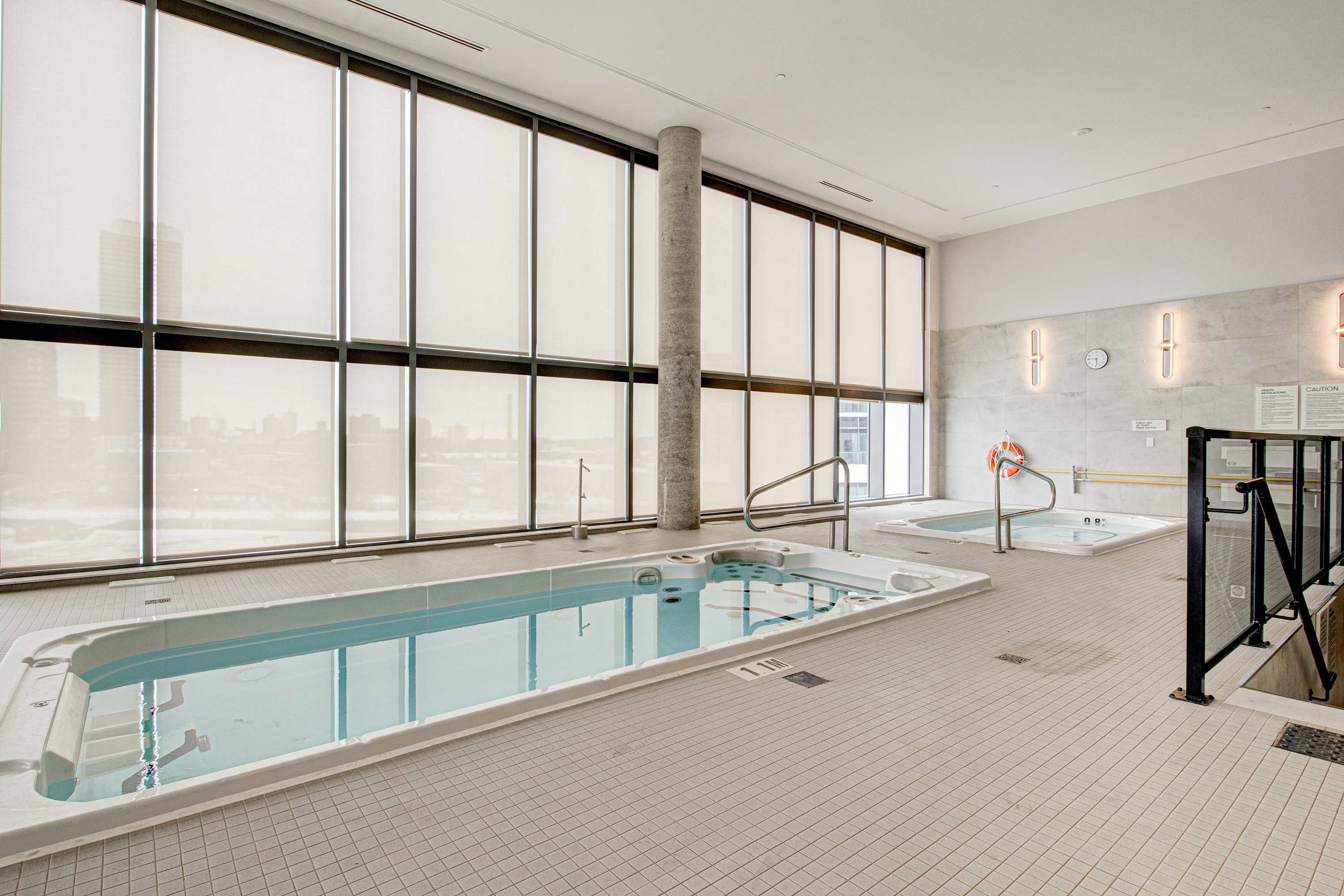 An indoor pool area features a long lap pool and two smaller hot tubs, all surrounded by light grey tiled flooring and large floor-to-ceiling windows with white blinds offering city views.