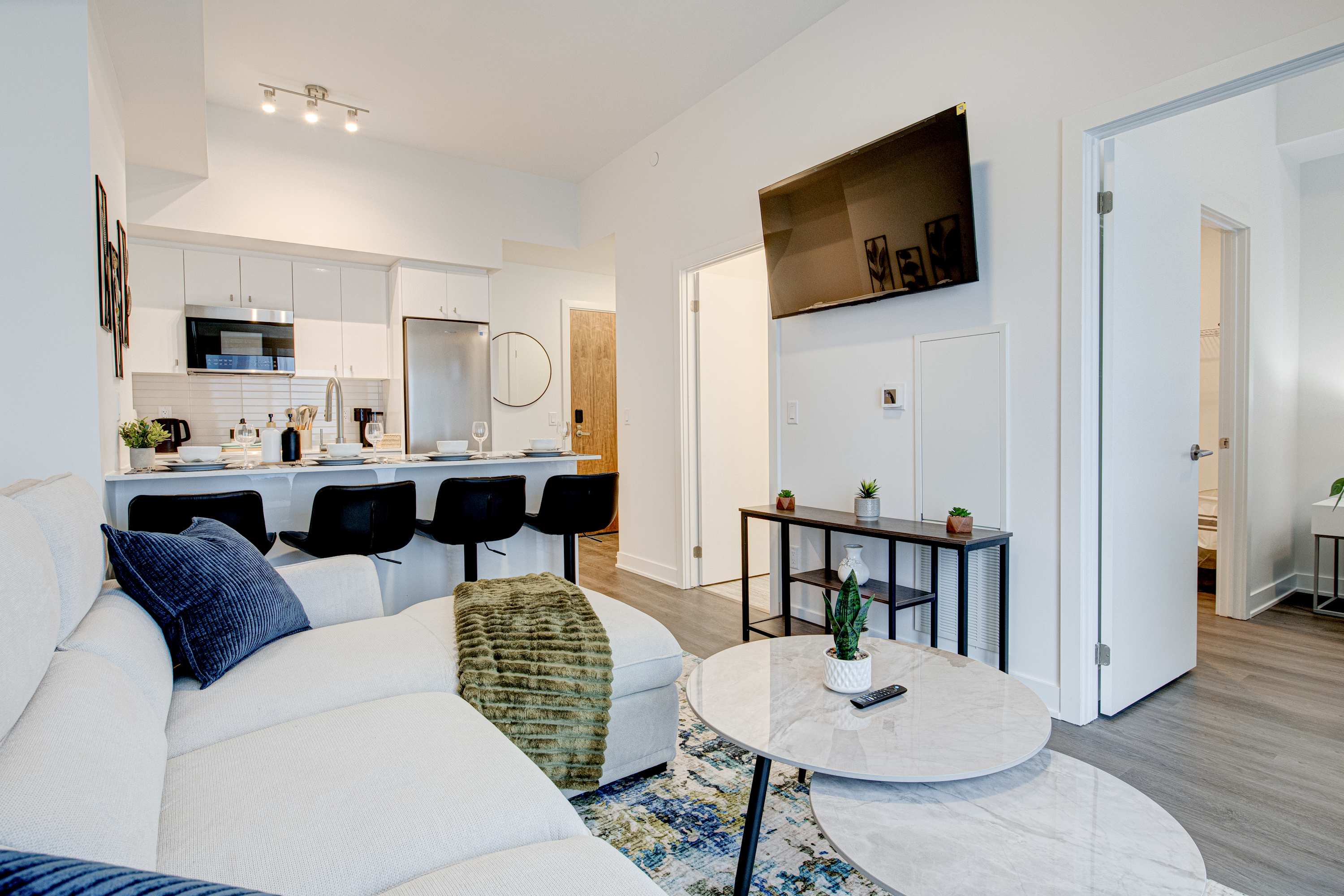 An open-concept living area features a light-colored sectional sofa facing a wall-mounted TV. A kitchen with white cabinetry and stainless steel appliances is visible in the background, along with a breakfast bar.