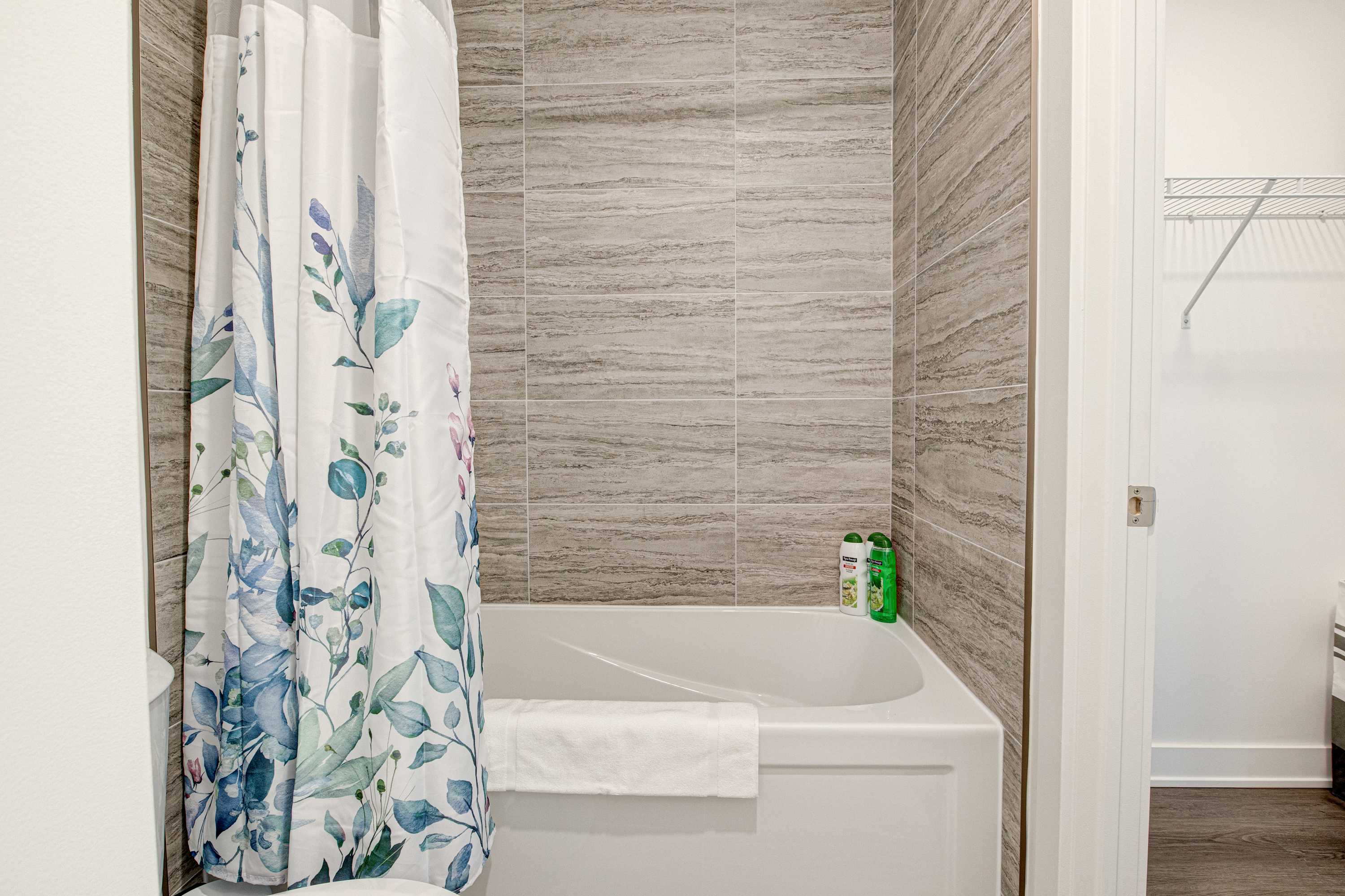 Clean bathtub with grey horizontal tiled walls and a white shower curtain featuring a blue and green floral pattern, with toiletries visible on the tub ledge.