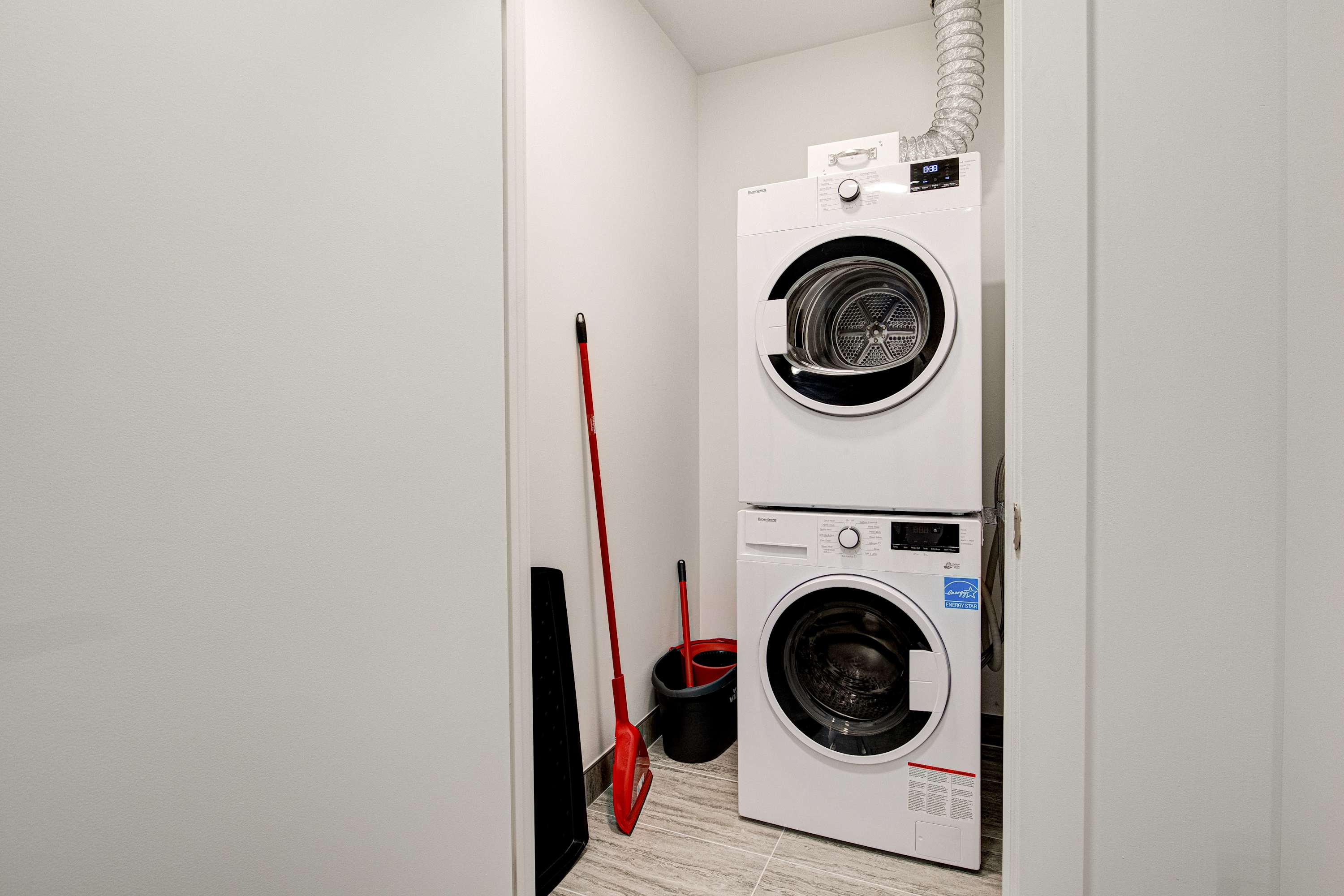 A compact laundry closet features a stacked white washer and dryer unit, with a red broom and dustpan leaning against the wall next to a black bucket, all on light grey tiled flooring.