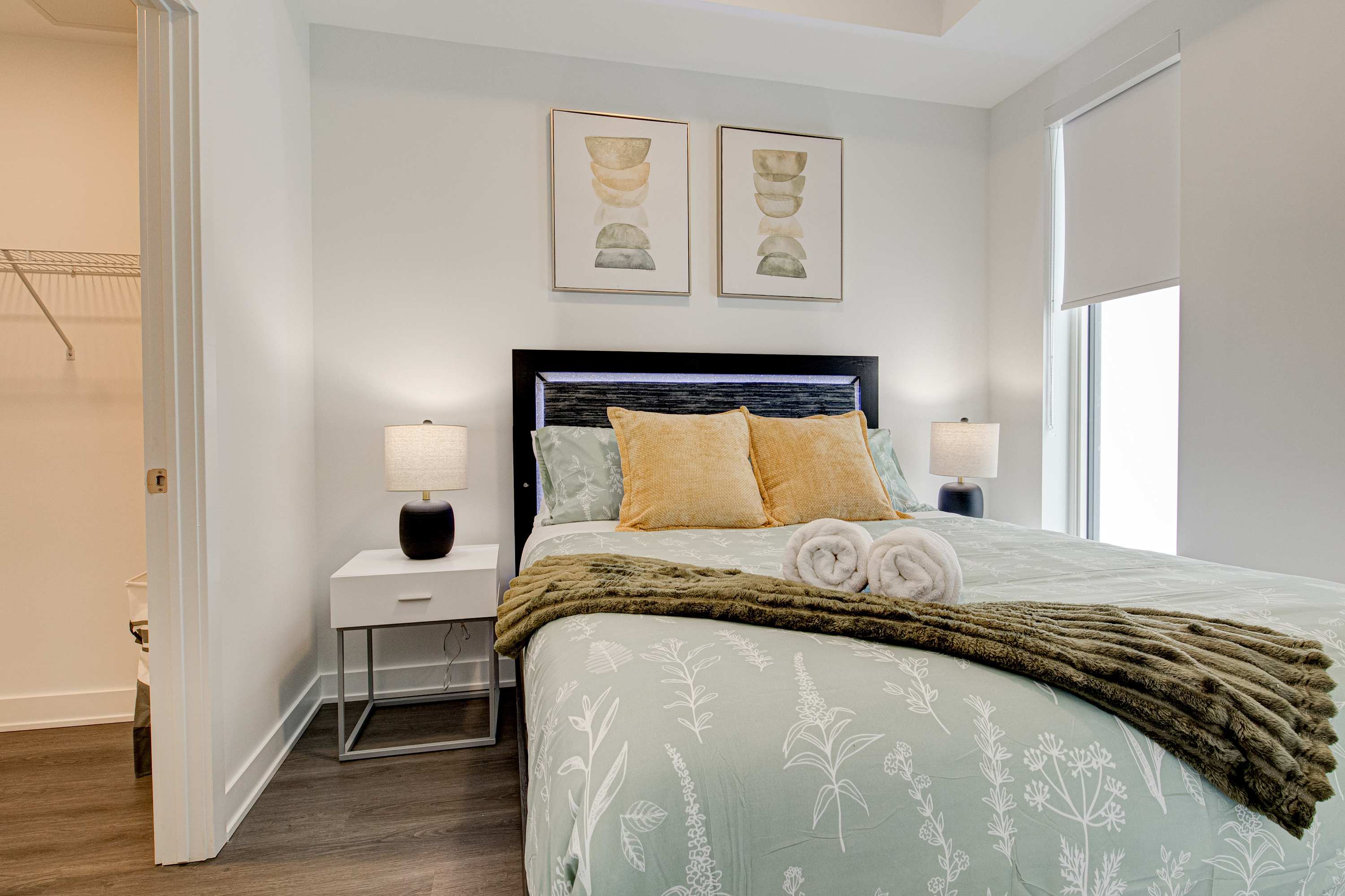 Well-lit bedroom featuring a queen-sized bed with light green and yellow bedding, flanked by white nightstands and lamps, with abstract art above the headboard and an open closet door revealing wire shelving.