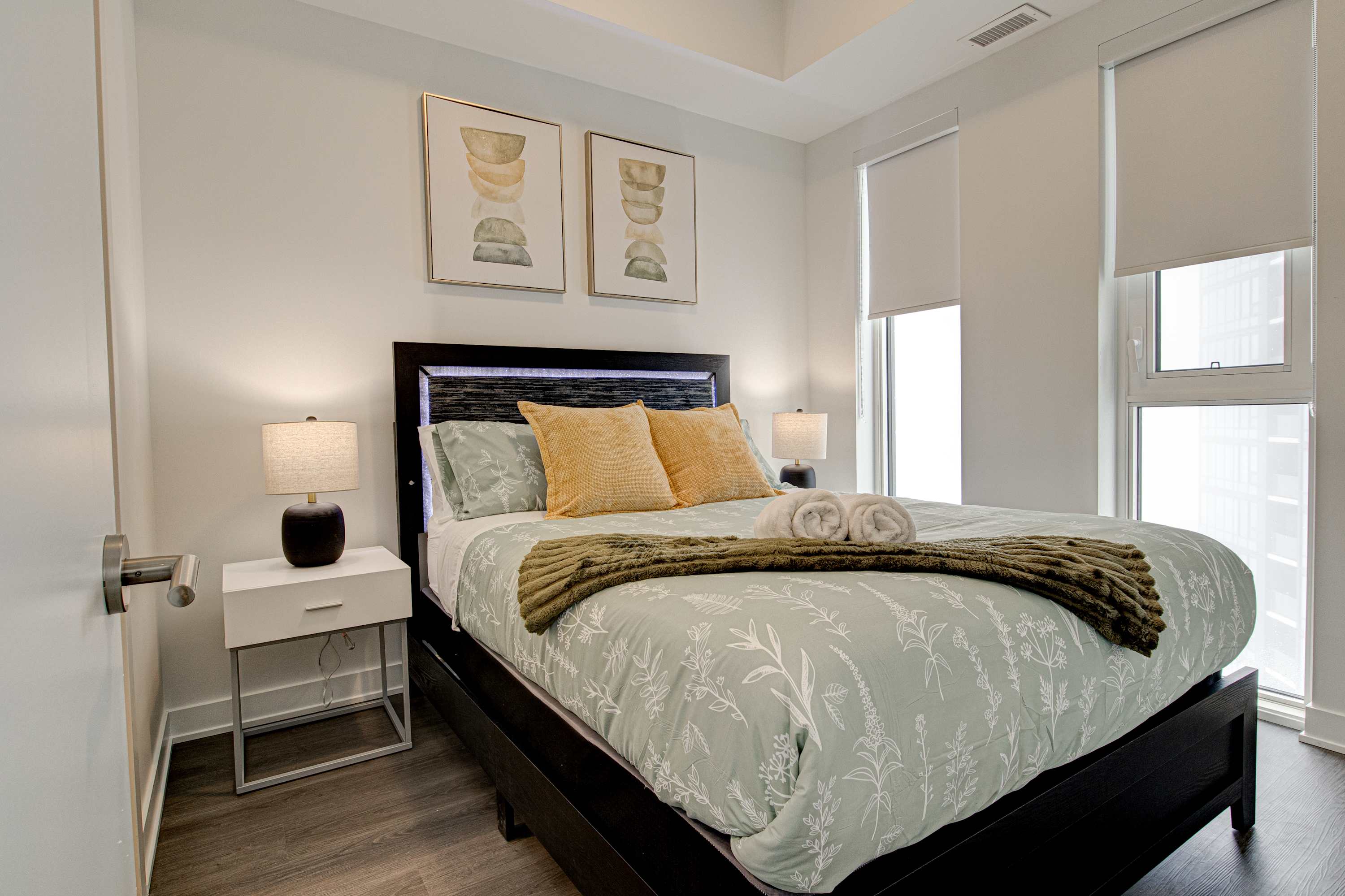 Cozy bedroom with a dark wood bed frame, light green and yellow bedding, two white nightstands with lamps, and abstract artwork above the headboard, with large windows providing natural light.