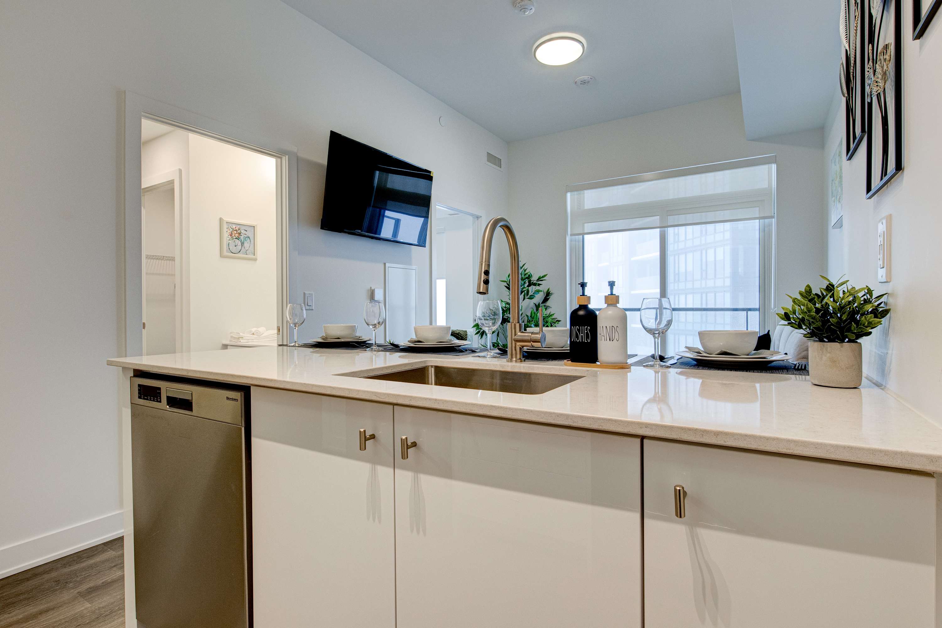 Modern kitchen island with a stainless steel sink and faucet, integrated dishwasher, and white cabinetry, featuring a breakfast bar with black stools and a wall-mounted TV in the background.