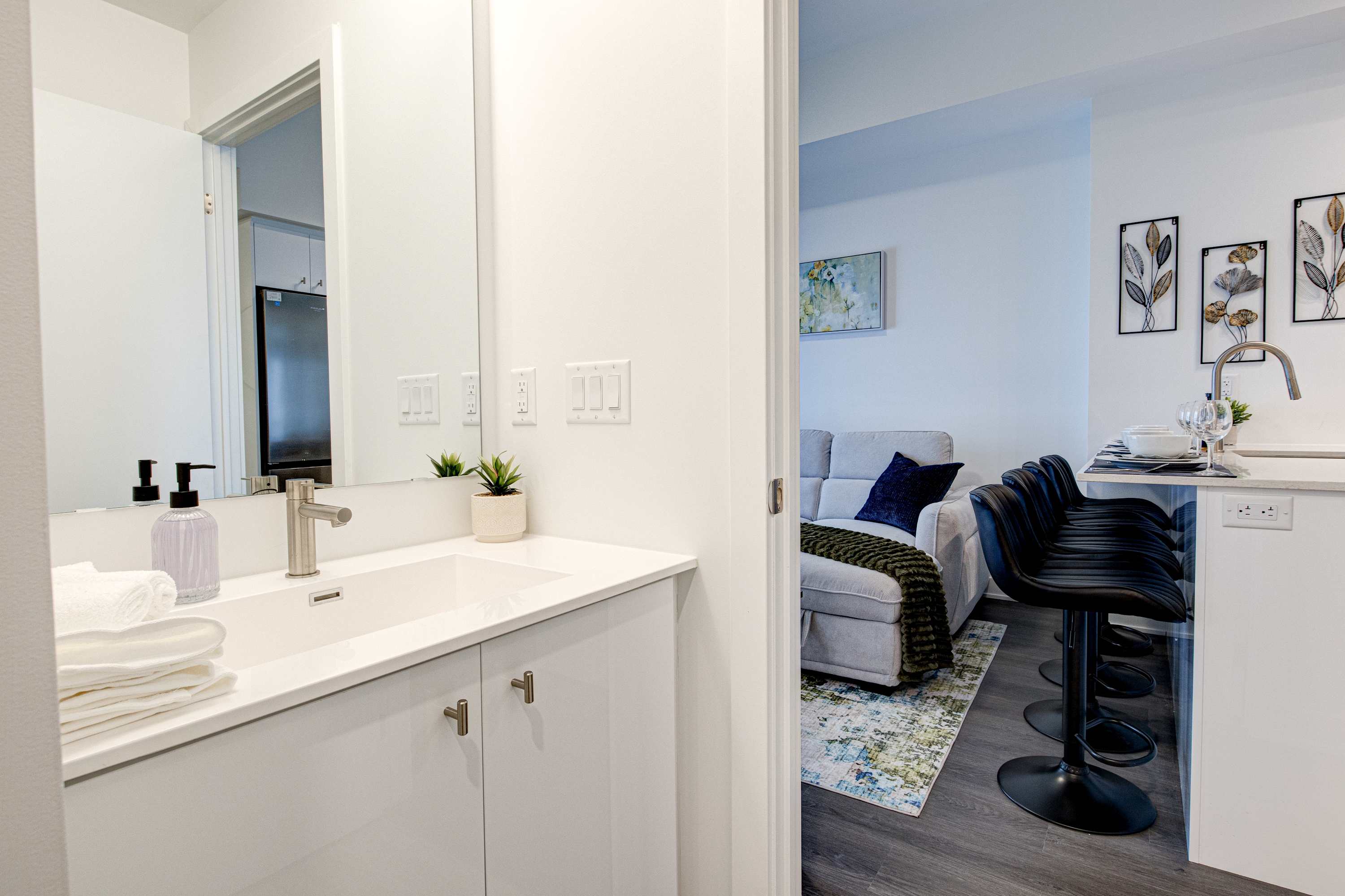 Bright bathroom featuring a white vanity with a sink and large mirror, looking out towards a living area with a grey sofa and a breakfast bar with black stools.