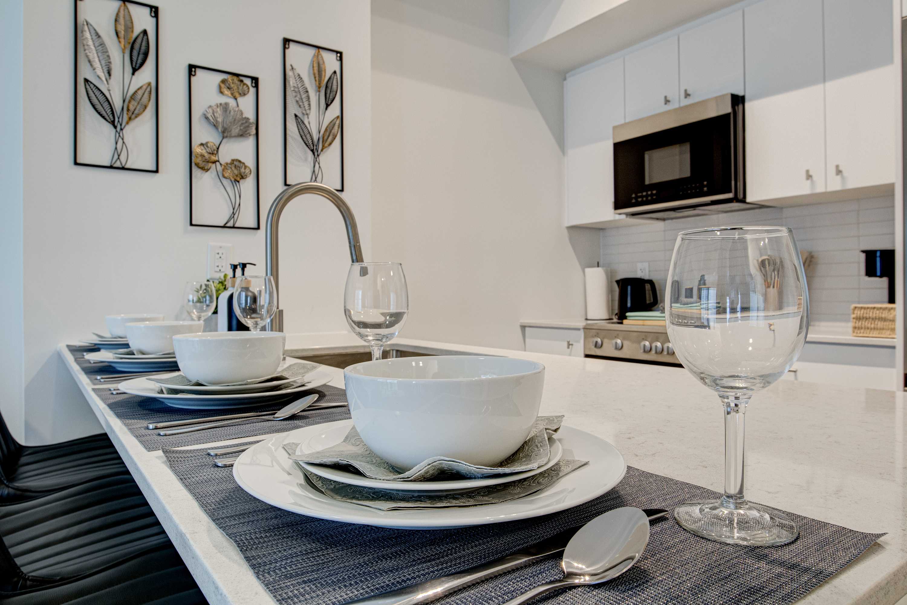 A close-up view of the kitchen island shows a set table with white bowls, plates, and wine glasses, ready for dining. A modern stainless steel faucet and a small plant are also visible on the white quartz countertop.