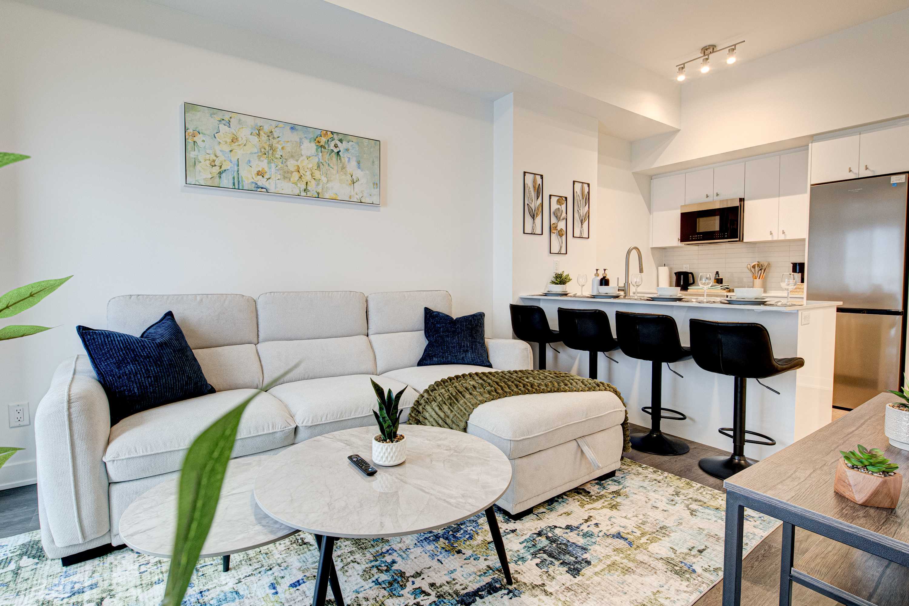 An open-concept living room with a light-colored sectional sofa and a colorful rug transitions into a modern kitchen with white cabinets and a breakfast bar. A large window provides natural light and a city view.