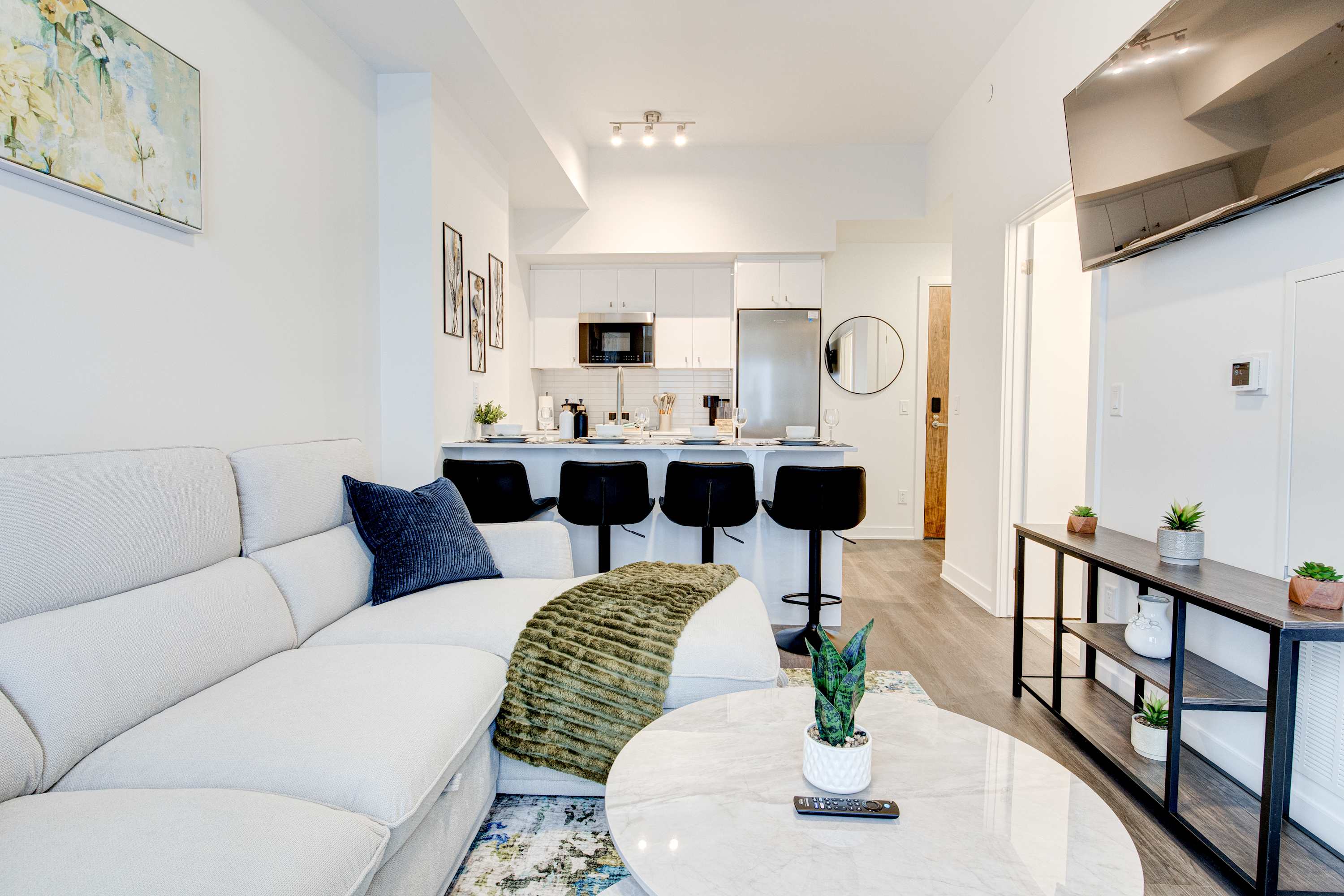 The open-concept living room features a light-colored sectional sofa with a green throw blanket and a marble coffee table. The adjacent kitchen has white cabinetry, stainless steel appliances, and a breakfast bar with black stools.